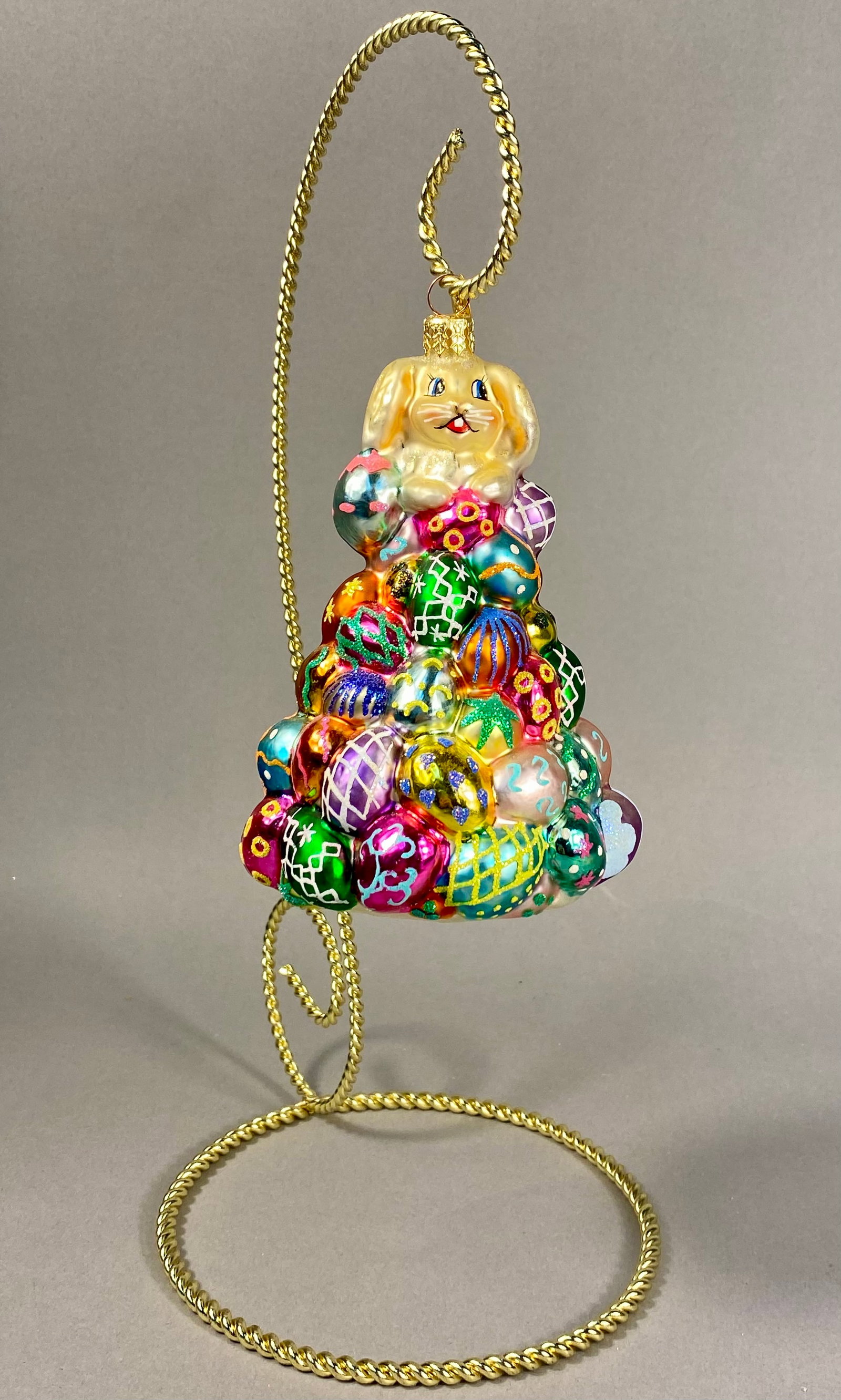 Radko Easter Bunny and Egg Pyramid Ornament: **Stand Not Included** Christopher Radko hand-blown glass holiday ornament featuring a cream-colored bunny with gold glitter accents sitting atop a pyramid-shaped pile of Easter eggs. Decoration inclu