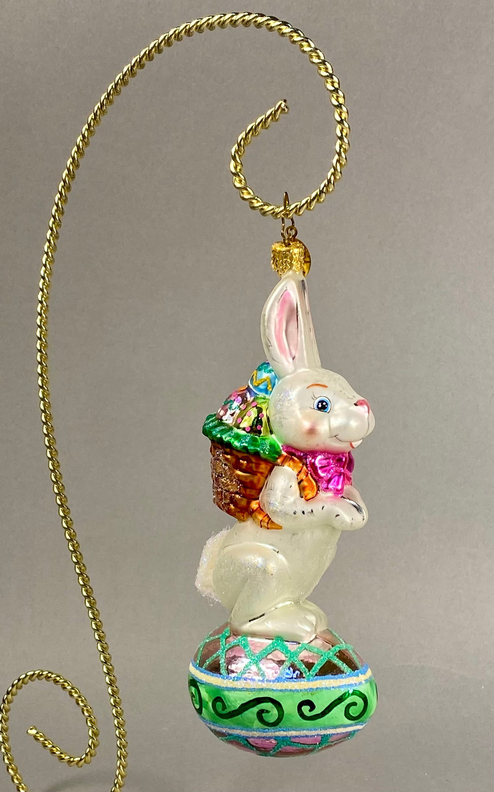 Radko Bunny Trail Basket Egg Blown Glass Ornament: **Stand Not Included** This is an authentic Christopher Radko blown glass holiday ornament depicting a white anthropomorphic rabbit standing on a large decorated Easter egg. The item is crafted from f