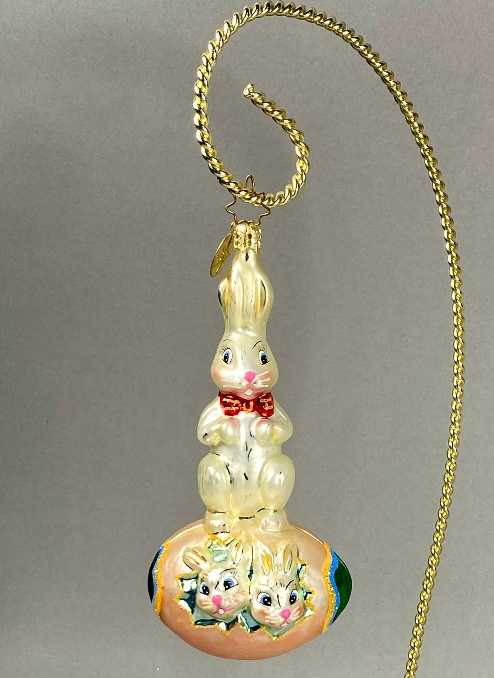 Radko Blown Glass Easter Bunnies in Egg Ornament: **Stand Not Included** This mouth-blown, silvered glass holiday ornament is a creation by Christopher Radko. The piece features a standing cream-colored rabbit wearing a red bow tie with gold-glittere