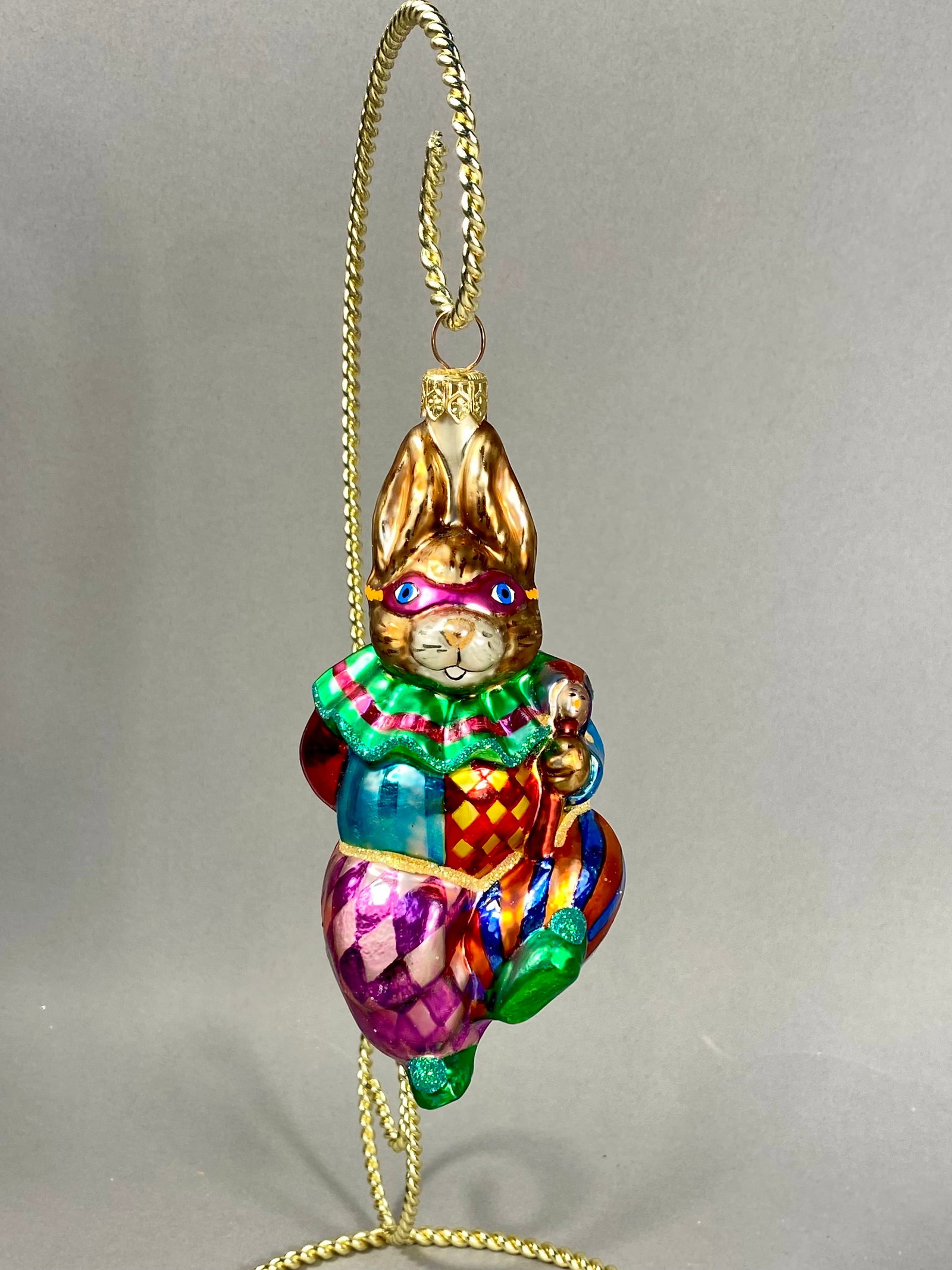 Radko Mask-Hare-Ade Rabbit Jester Glass Ornament: **Stand Not Included** A mouth-blown glass ornament from the Christopher Radko Mask-Hare-Ade collection. The ornament depicts a rabbit dressed in a vibrant harlequin or court jester costume. The rabbi
