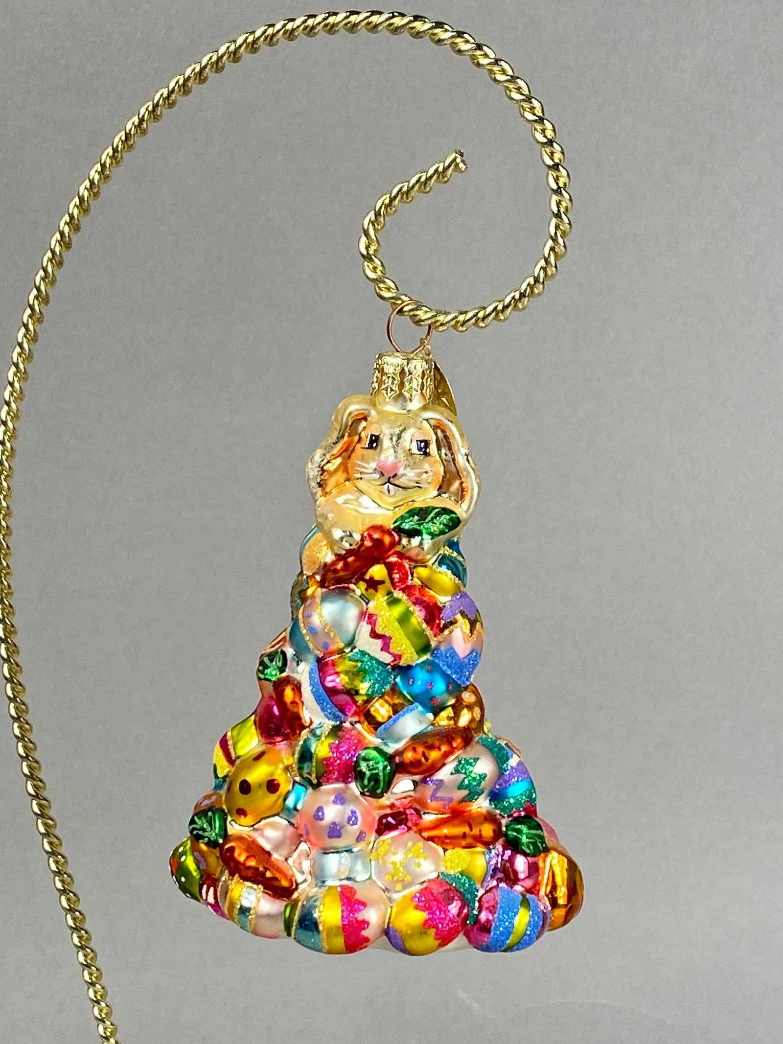 Christopher Radko Easter Rabbit and Egg Tree Glass Ornament: A Christopher Radko mouth-blown glass figural ornament featuring a light gold-colored rabbit seated atop a conical stack of Easter-themed elements. The base of the figure is a dense arrangement of mul