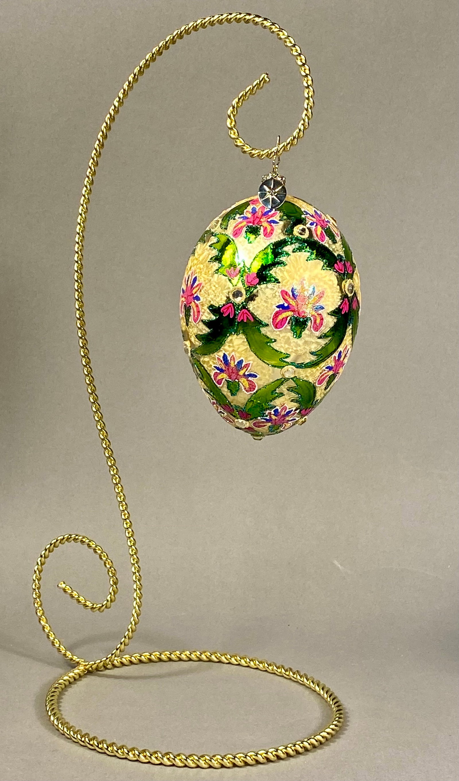 Christopher Radko Hand-Painted Blown Glass Faberge-Style Egg Ornament on Stand: A hand-painted blown-glass egg-shaped ornament produced by Christopher Radko. The ornament features an intricate floral design with stylized blossoms in pink, blue, and yellow, interspersed with green