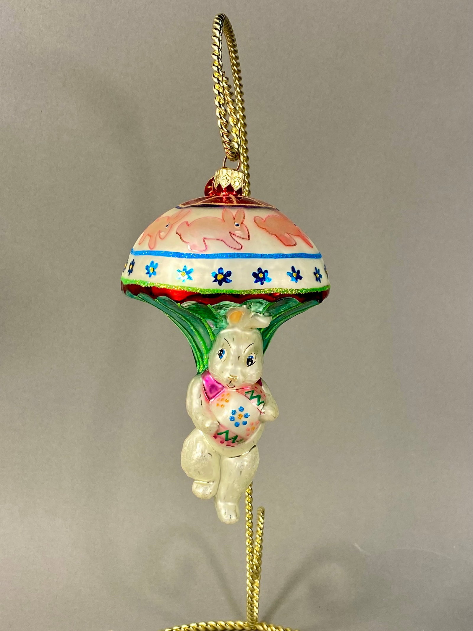 Christopher Radko Bunnies Away Parachuting Rabbit Glass Ornament: A Christopher Radko hand-blown glass holiday ornament titled Bunnies Away. The piece features a white rabbit character depicted in a parachuting pose while clutching a decorated Easter egg. The egg is