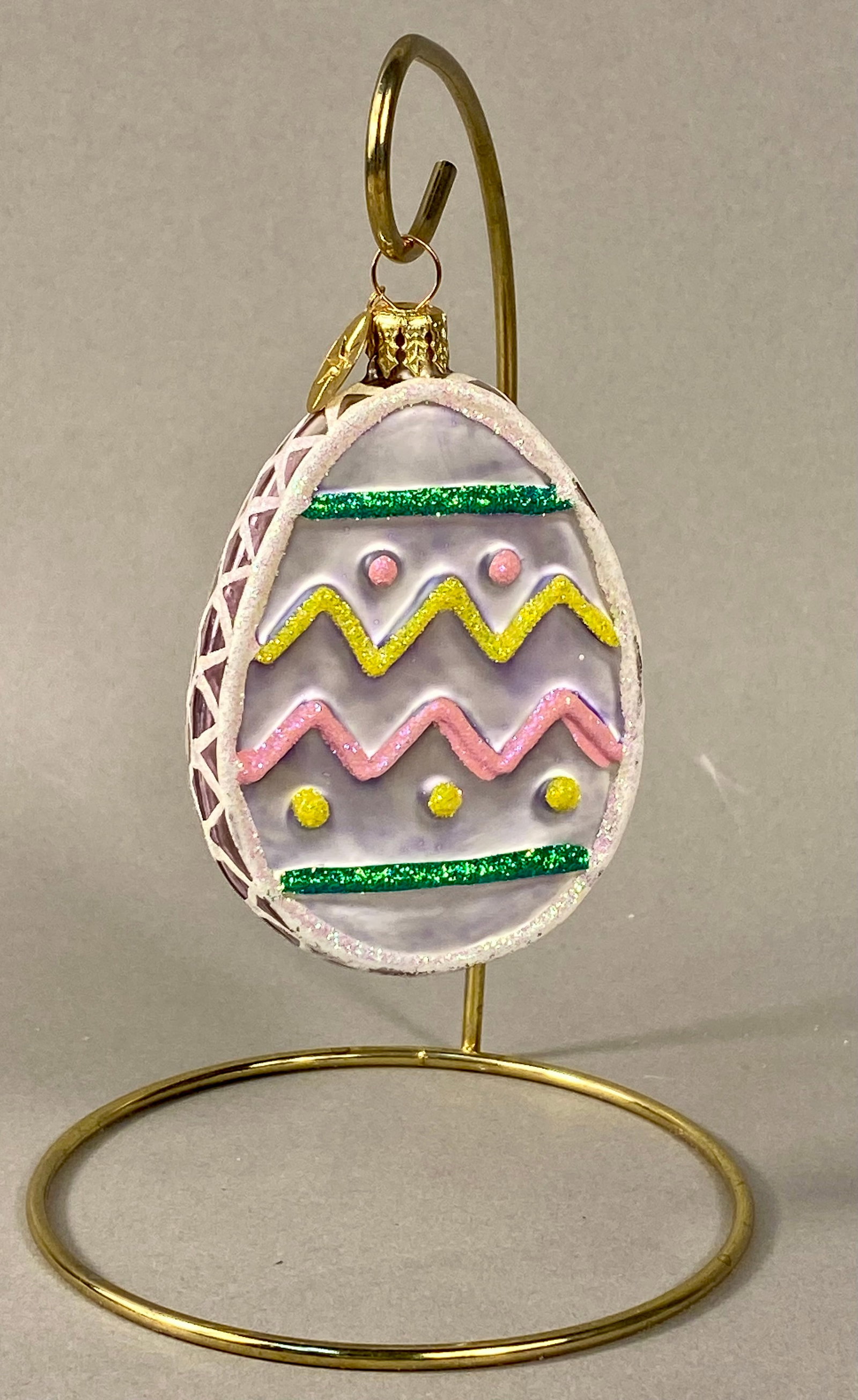 Christopher Radko Mouth-Blown Glass Easter Egg Ornament: A mouth-blown glass Easter egg ornament produced by Christopher Radko. The ornament features an iridescent lavender glass body with a central decorative panel framed by a raised, textured white glitte