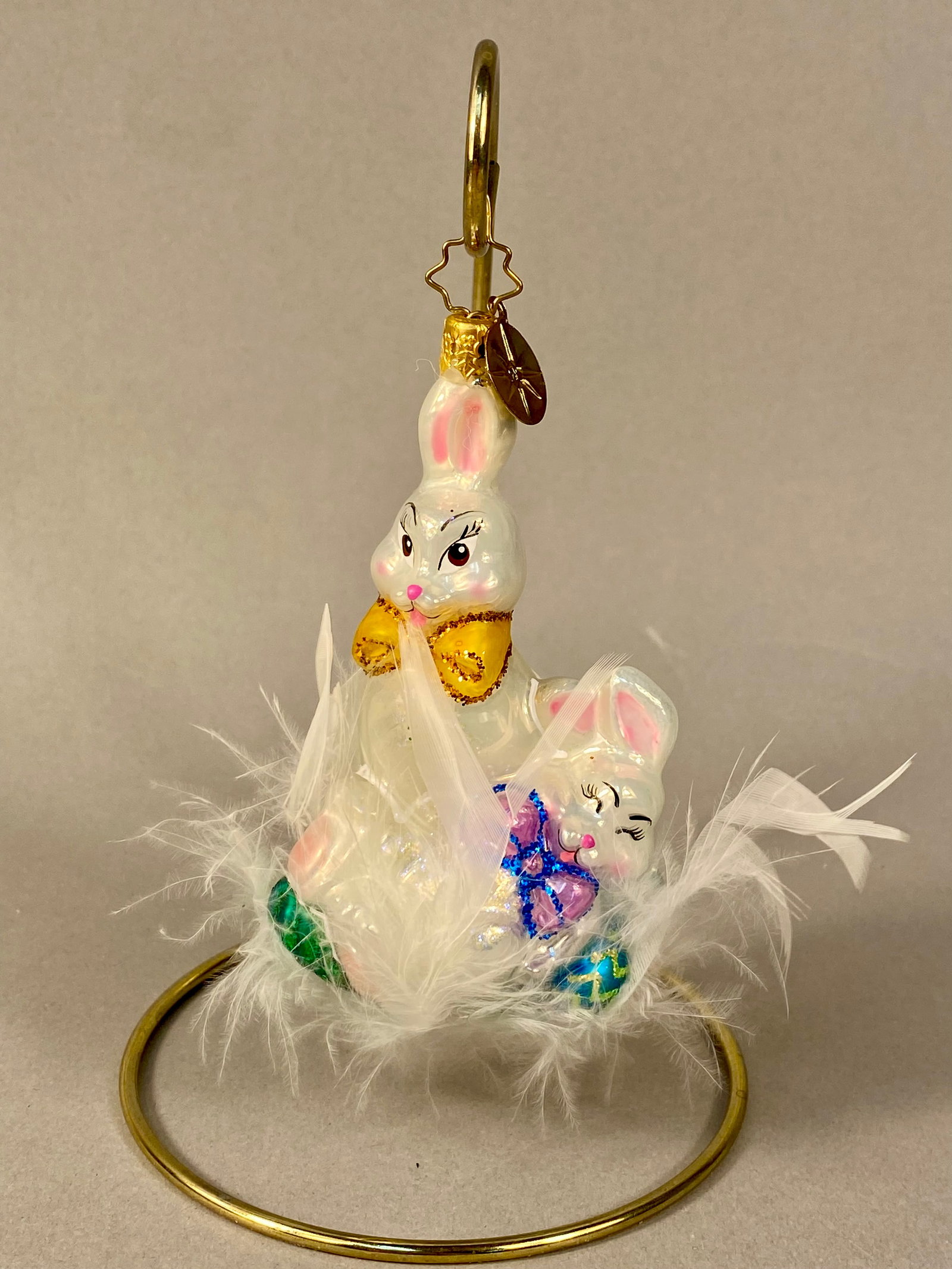 Christopher Radko Hoppy Holidays Glass Easter Bunny Ornament: This hand-blown glass ornament, titled Hoppy Holidays, is produced by Christopher Radko. The piece features two white rabbits with a pearlescent finish, hand-painted details, and glitter accents nestl