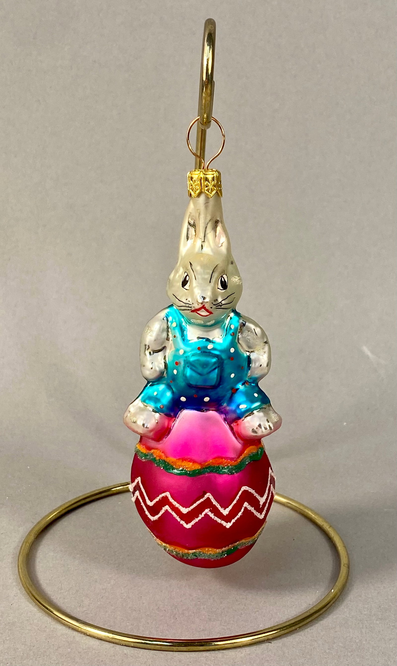 Vintage Czechoslovakian Hand-Blown Glass Rabbit on Egg Christmas Ornament: This vintage hand-blown glass ornament depicts a white rabbit perched on a decorated fuchsia pink egg. The rabbit is shown wearing bright blue overalls embellished with hand-painted white and red polk