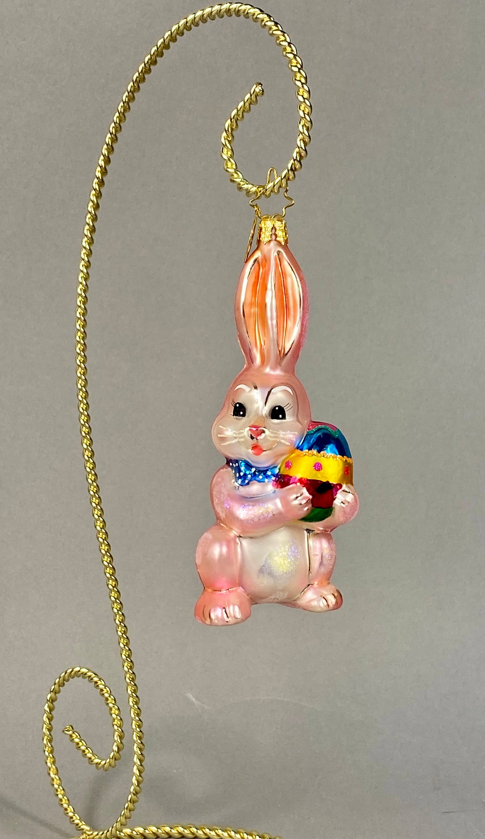 Christopher Radko Hoppy Holidays Hand Blown Glass Easter Bunny Ornament: This Christopher Radko hand-blown glass holiday ornament is titled Hoppy Holidays and features a standing rabbit with an iridescent pink pearlescent finish. The bunny is depicted holding a large Easte