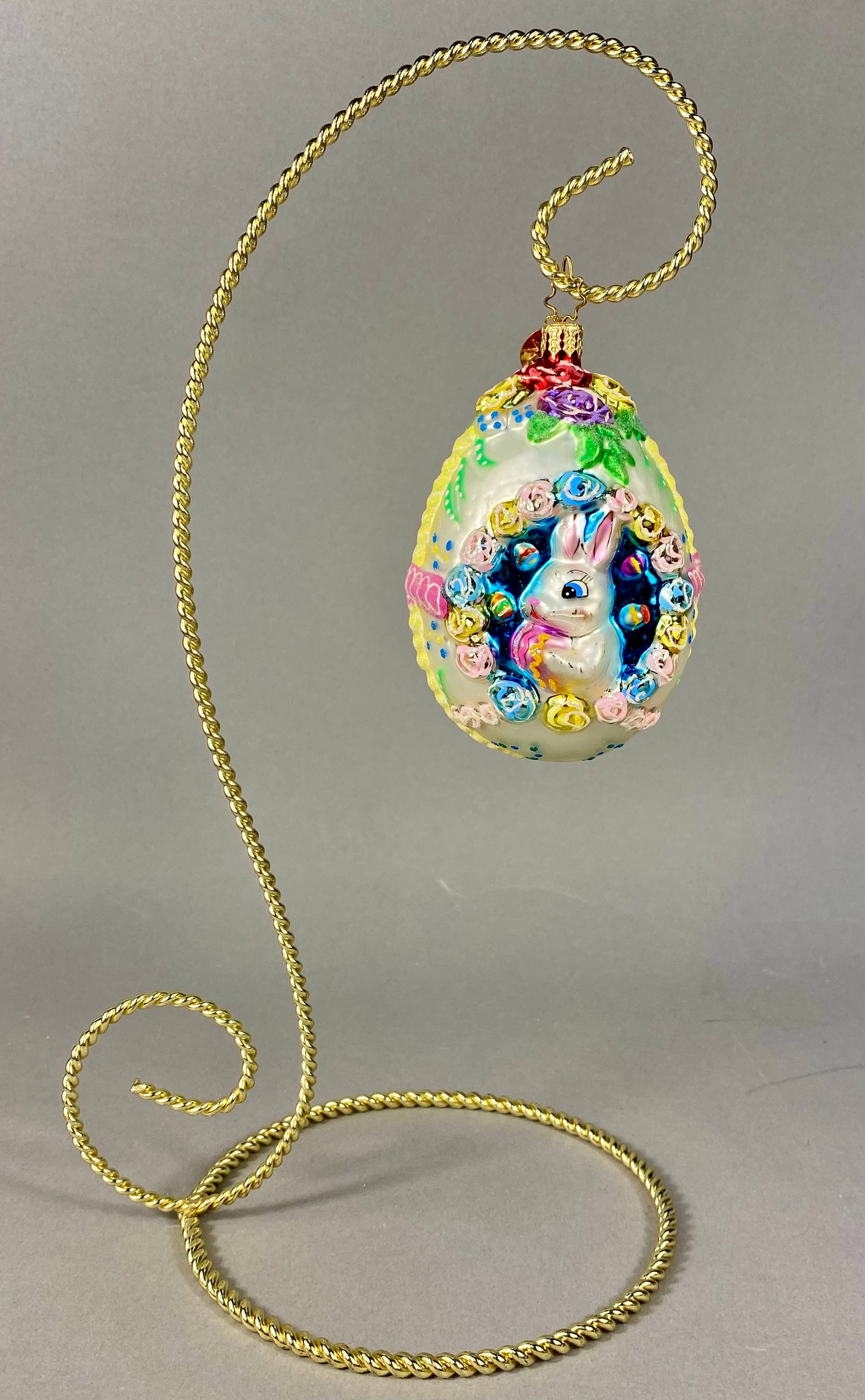 Christopher Radko Mouth-Blown Glass Bunny Easter Egg Ornament on Stand: A mouth-blown and hand-painted glass Easter ornament produced by Christopher Radko. The egg-shaped ornament features an iridescent white ground with a central recessed reflector style scene. This scen