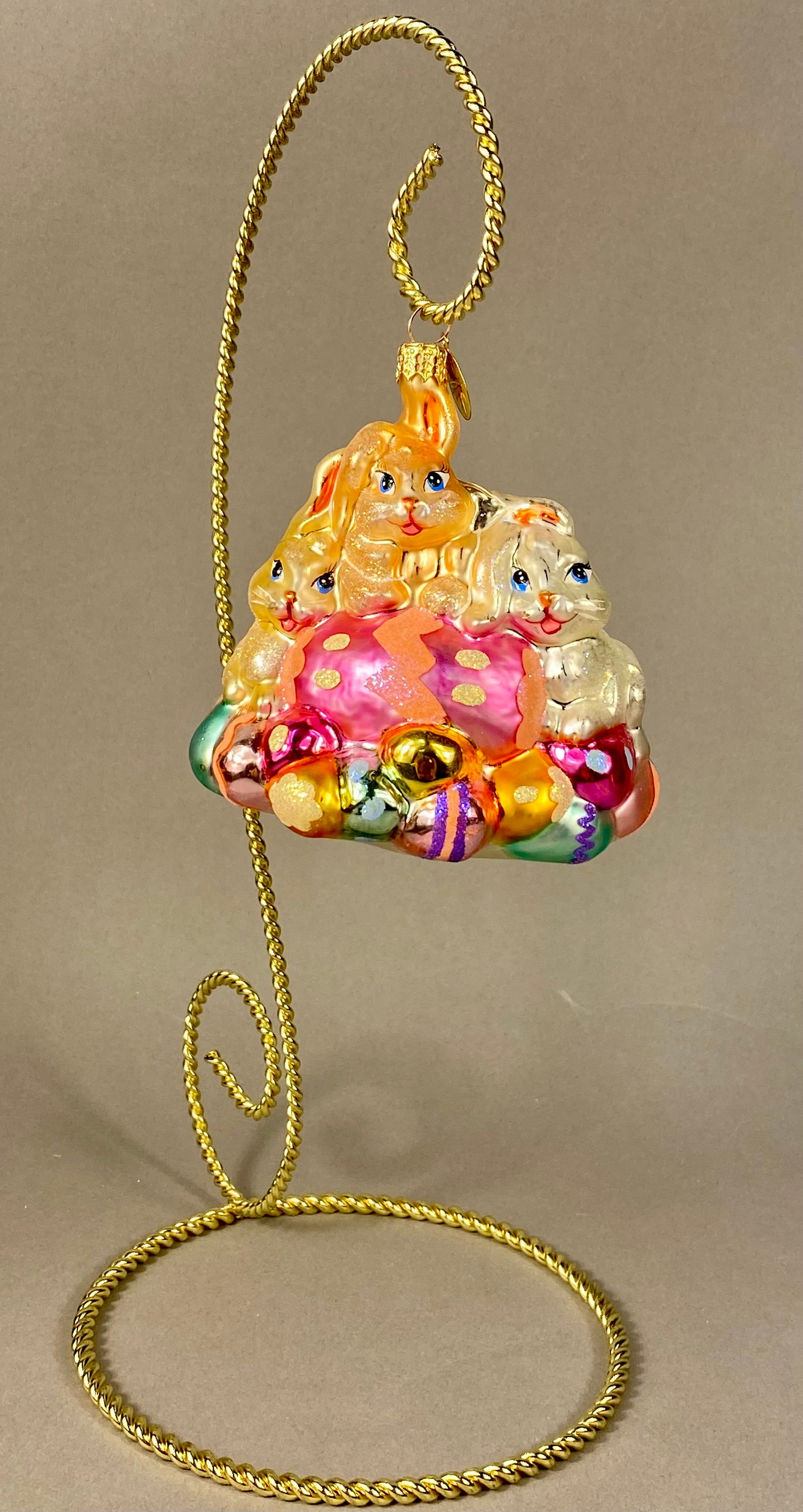 Christopher Radko Hand-Blown Glass Easter Bunnies and Eggs Holiday Ornament: A mouth-blown glass holiday ornament produced by Christopher Radko, featuring an Easter theme. The composition consists of a trio of rabbits perched upon a large, magenta-colored egg. The central rabb
