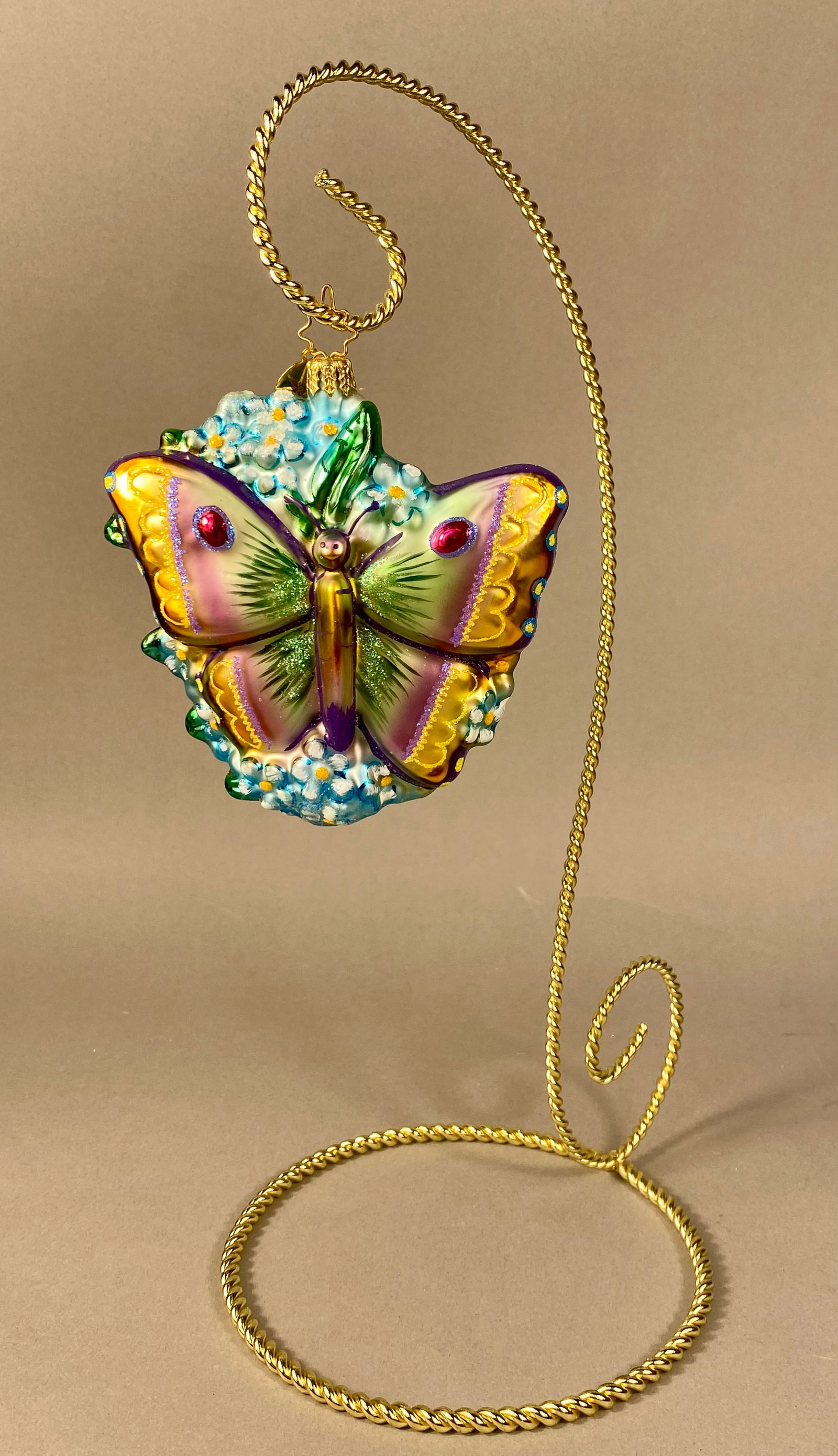Christopher Radko Hand-Blown Glass Butterfly and Forget-Me-Not Ornament: A hand-blown glass ornament from Christopher Radko, featuring a stylized butterfly perched atop a cluster of light blue forget-me-not flowers. The butterfly is detailed with hand-painted lacquer in sh
