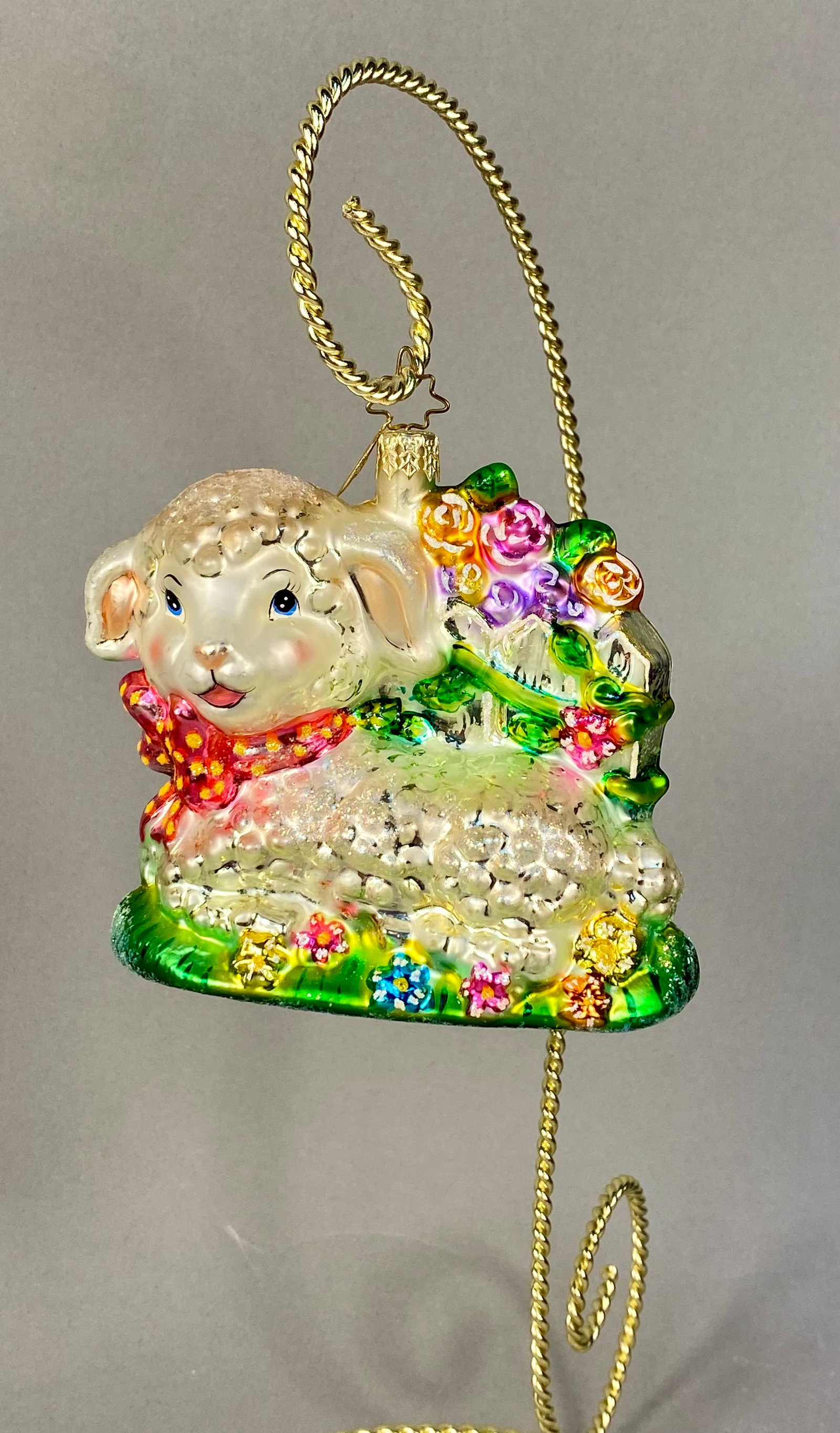 Figural Blown Glass Lamb Christmas Ornament with Picket Fence and Roses: This mouth-blown silvered glass Christmas ornament features a figural design of a seated white lamb with a textured, pearlescent finish and iridescent glitter accents. The lamb wears a red bow tie wit