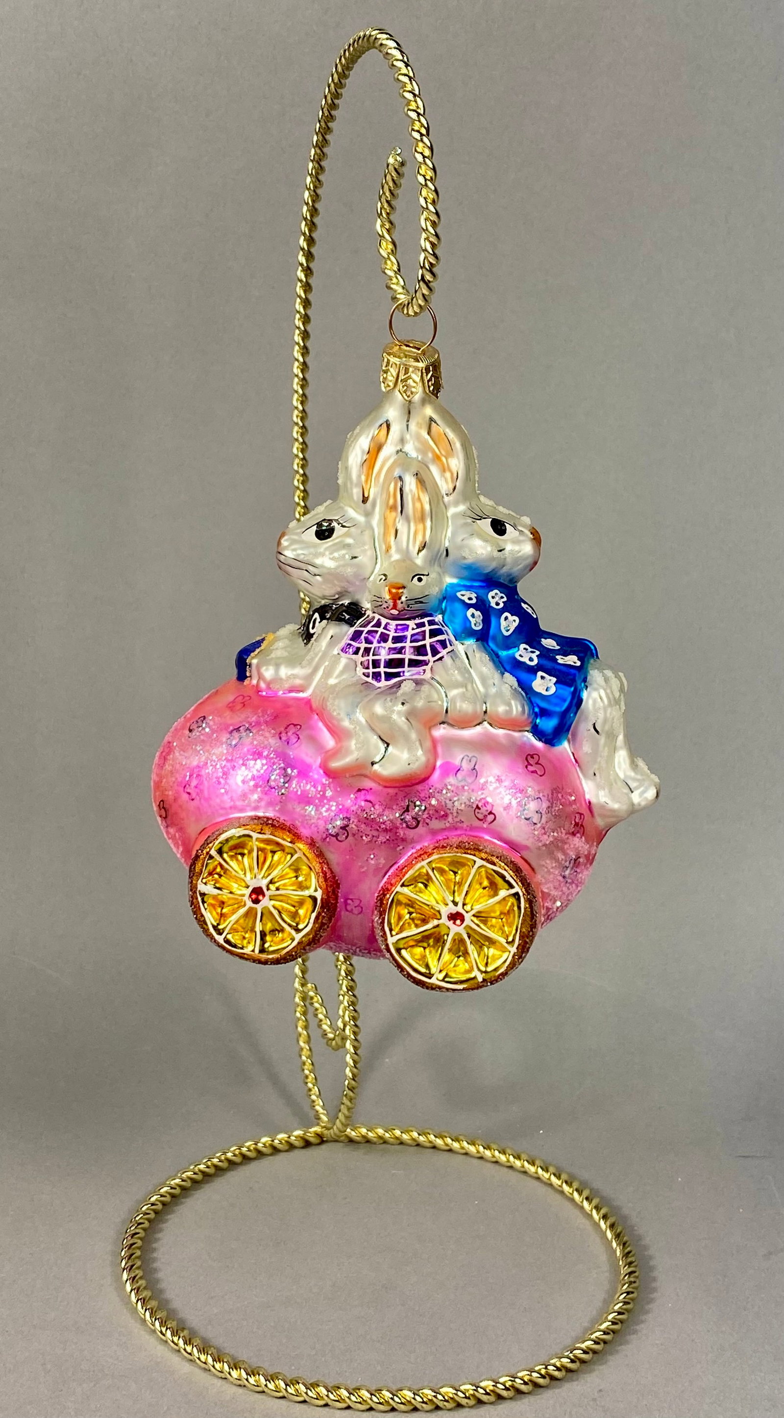 Christopher Radko Bunmobile Blown Glass Easter Bunny Egg Carriage Ornament: This hand-blown glass holiday ornament is a Christopher Radko design titled Bunmobile. The piece depicts three white rabbits seated atop a large, iridescent pink egg-shaped carriage. The carriage is e