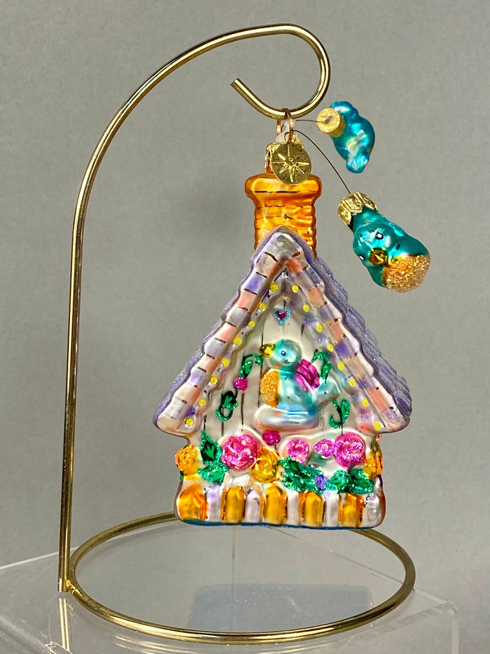 Christopher Radko Hand-Blown Glass Birdhouse Cottage Holiday Ornament: A mouth-blown glass holiday ornament by Christopher Radko, expertly crafted in the form of a whimsical birdhouse cottage. The front facade features a blue bird nestled within an arched niche, surround