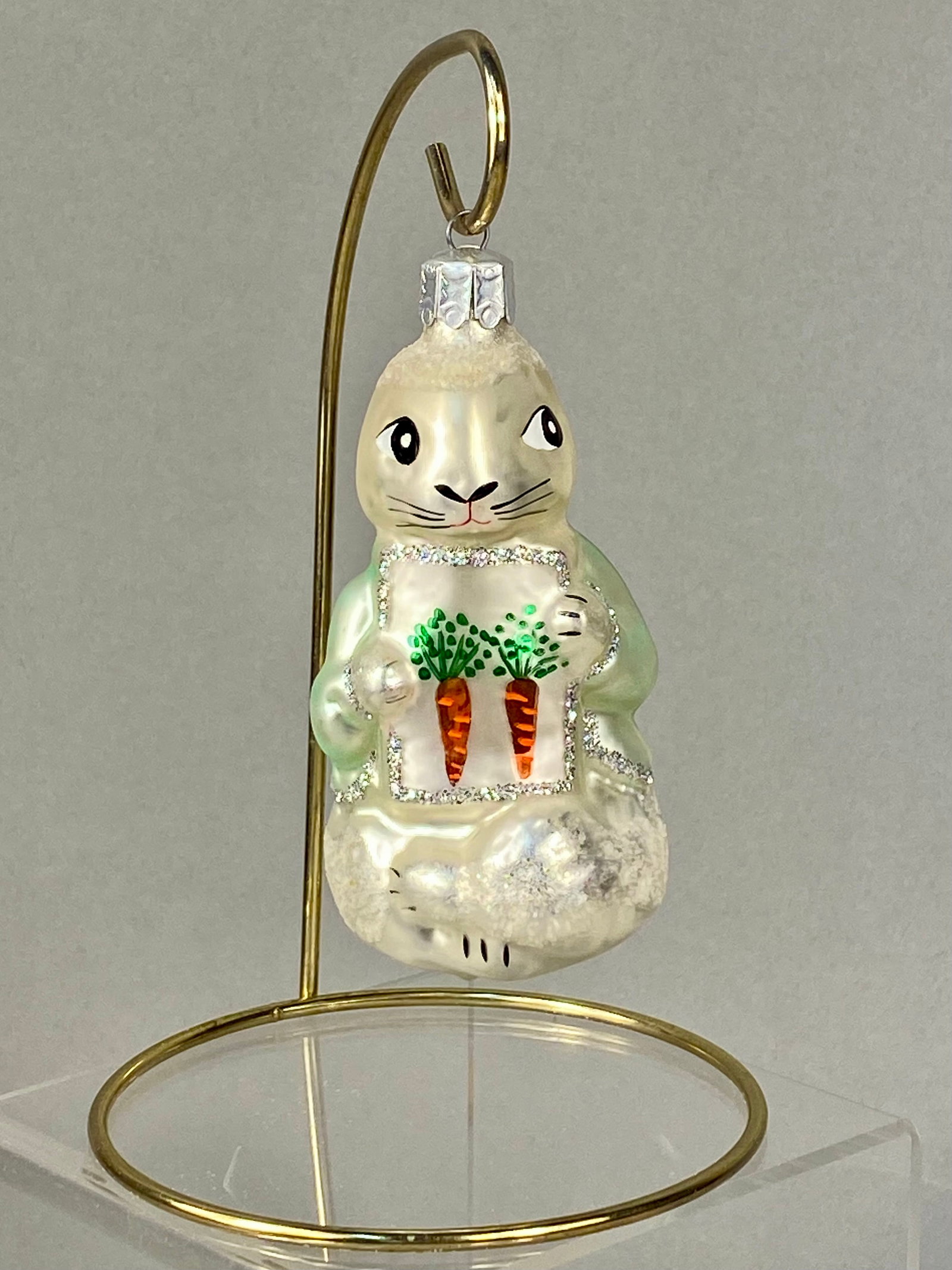 Christopher Radko Hand-Blown Glass Rabbit with Carrots Ornament: A hand-blown glass holiday ornament by Christopher Radko, featuring an upright standing rabbit. The figurine is finished in a pearlescent cream color and is depicted wearing a pale green jacket accent