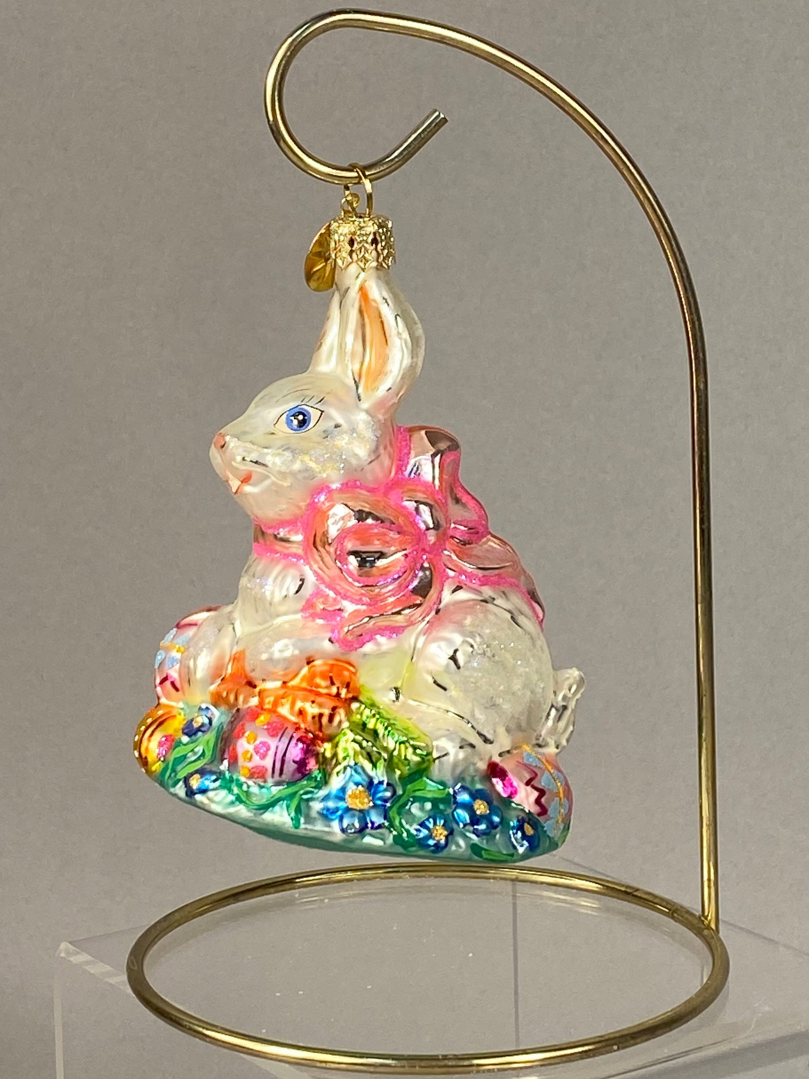 Christopher Radko Hoppy Holidays Glass Easter Bunny Ornament: A Christopher Radko mouth-blown glass seasonal ornament titled Hoppy Holidays. The piece depicts a seated white rabbit with blue eyes wearing a large iridescent pink bow around its neck. The rabbit is