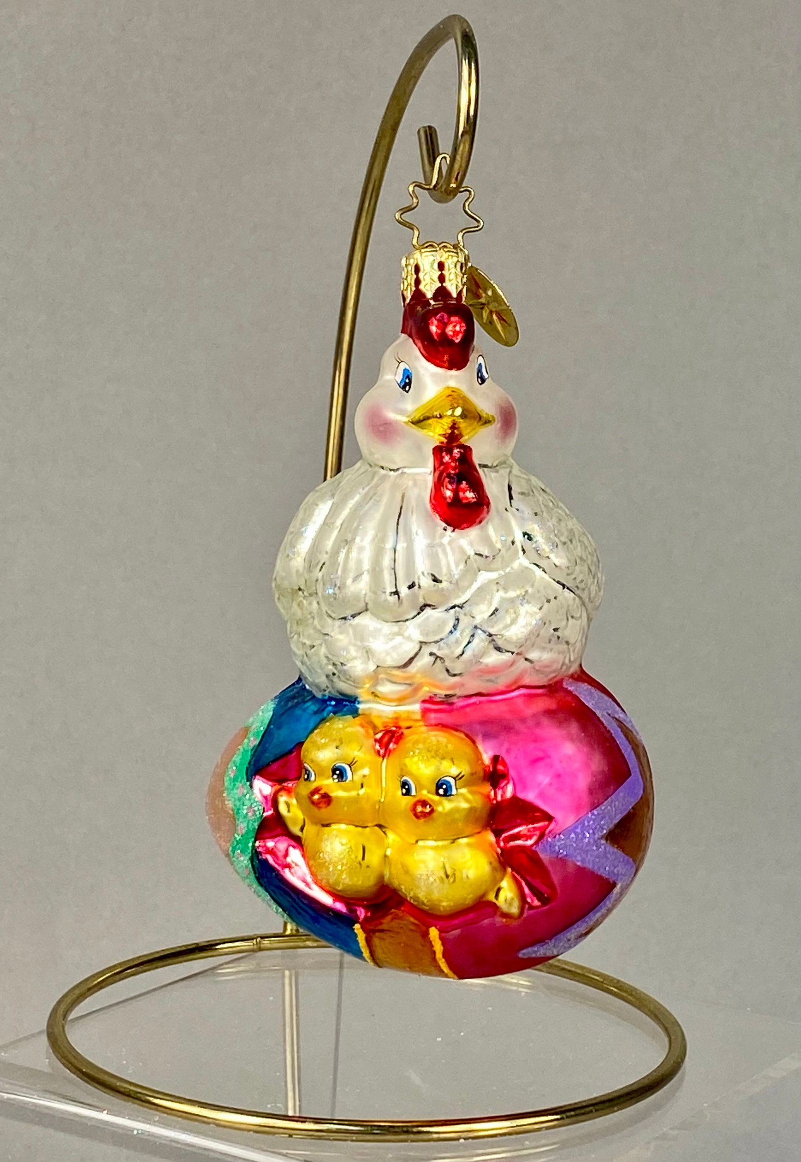 Christopher Radko Hand-Blown Glass Mother Hen on Easter Egg Ornament: A Christopher Radko mouth-blown and hand-painted glass ornament depicting a mother hen perched on a decorated Easter egg. The hen is rendered in pearlescent white glass with a red comb and wattle, yel