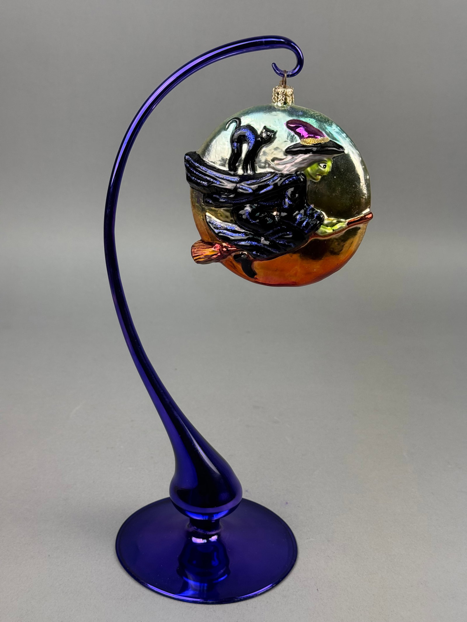 Radko Blown Glass Halloween Witch Ornament: This hand-painted blown glass ornament is designed as a flattened circular disc with an iridescent finish. The ornament is topped with a gold-toned metal cap embossed with an eight-pointed star, a sig