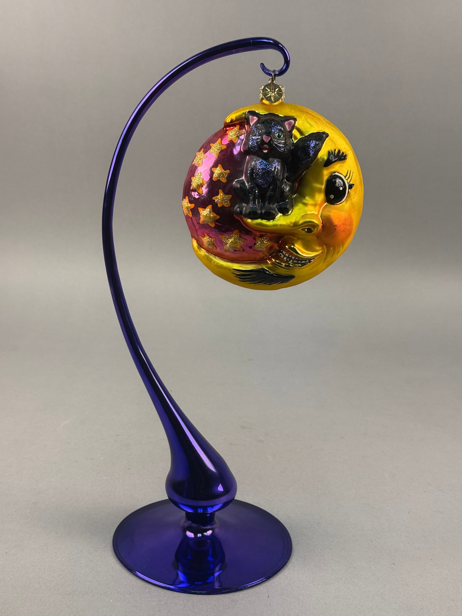 Radko Witchy Whiskers Halloween Glass Ornament: Christopher Radko Witchy Whiskers mouth-blown glass ornament. The design features a black cat with green eyes and blue glittered accents lounging on a pink iridescent orb adorned with raised, gold-gli