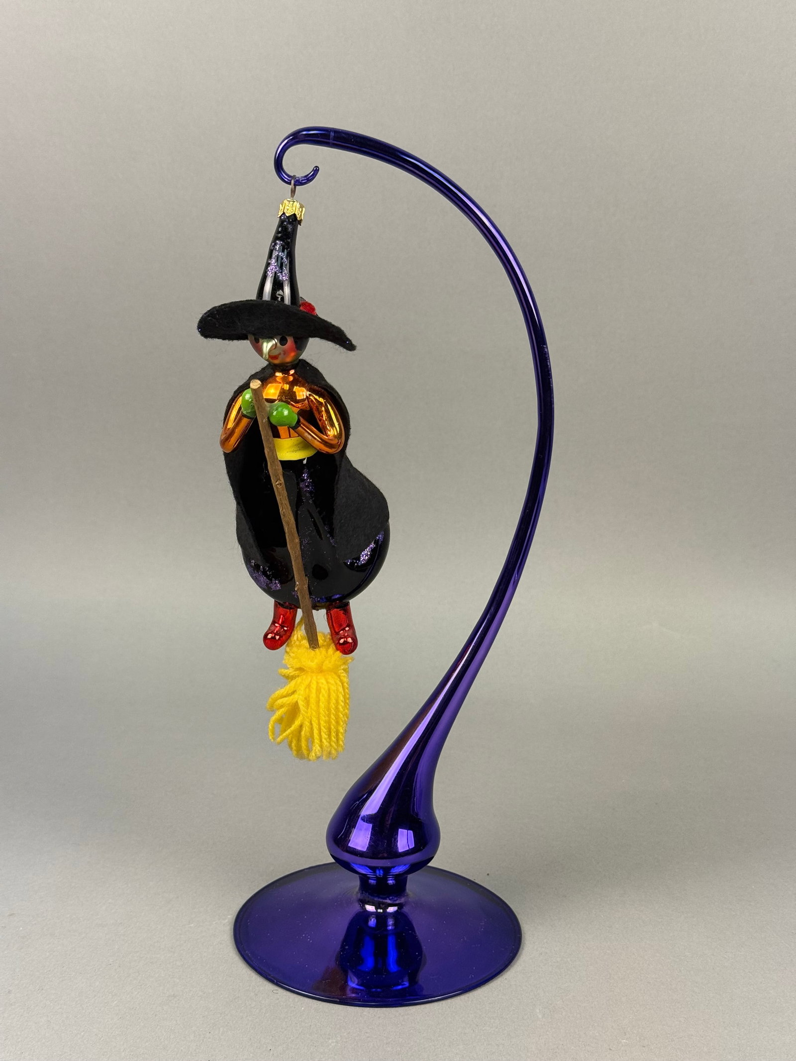 Radko Glass Witch on Broomstick Ornament: This blown glass ornament, produced by Christopher Radko depicts a whimsical witch in flight on a broomstick. The figure is decorated with various hand-painted and metallic finishes, featuring a black