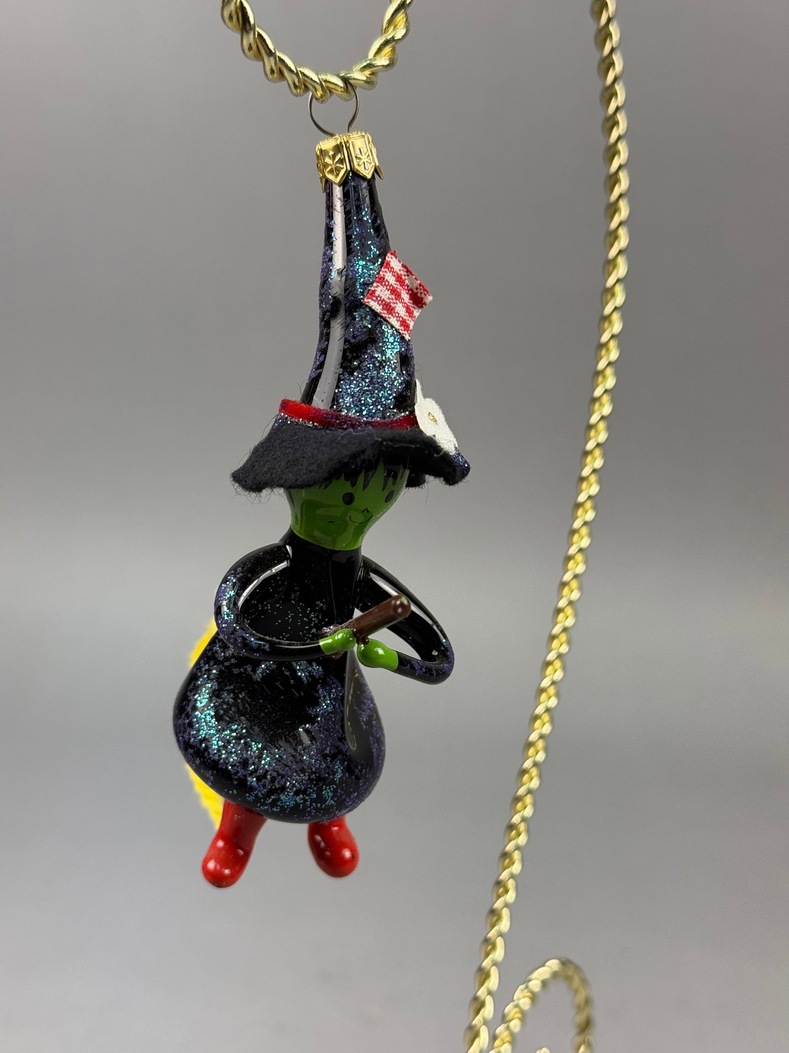 Radko Blown Glass Witch on Broomstick Ornament: A whimsical hand-blown glass ornament depicting a witch riding a broomstick. The figure is crafted with a dark glass body and hat finished in iridescent blue and green glitter. A decorative gold-tone 