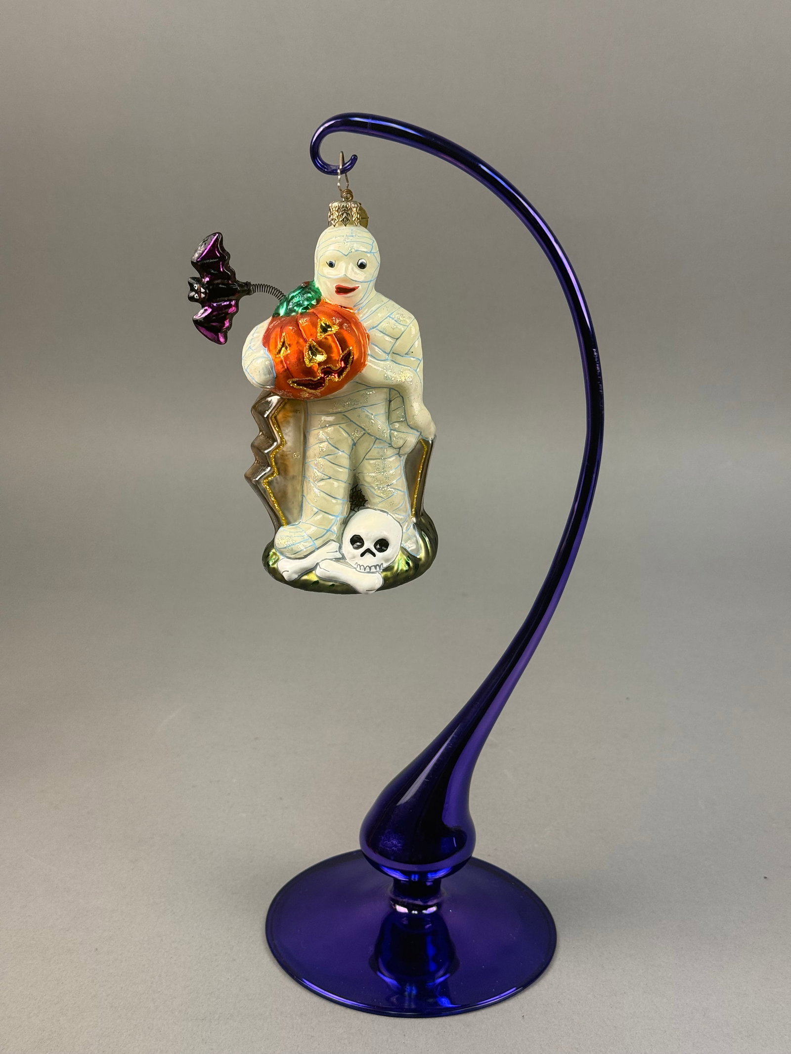 Radko Mummy in Coffin Glass Halloween Ornament: A Christopher Radko hand-painted blown glass ornament featuring a mummy in a coffin with a jack-o-lantern and dangling bat. Ornament stand NOT INCLUDED measuring approximately 5 inches in length. Exce