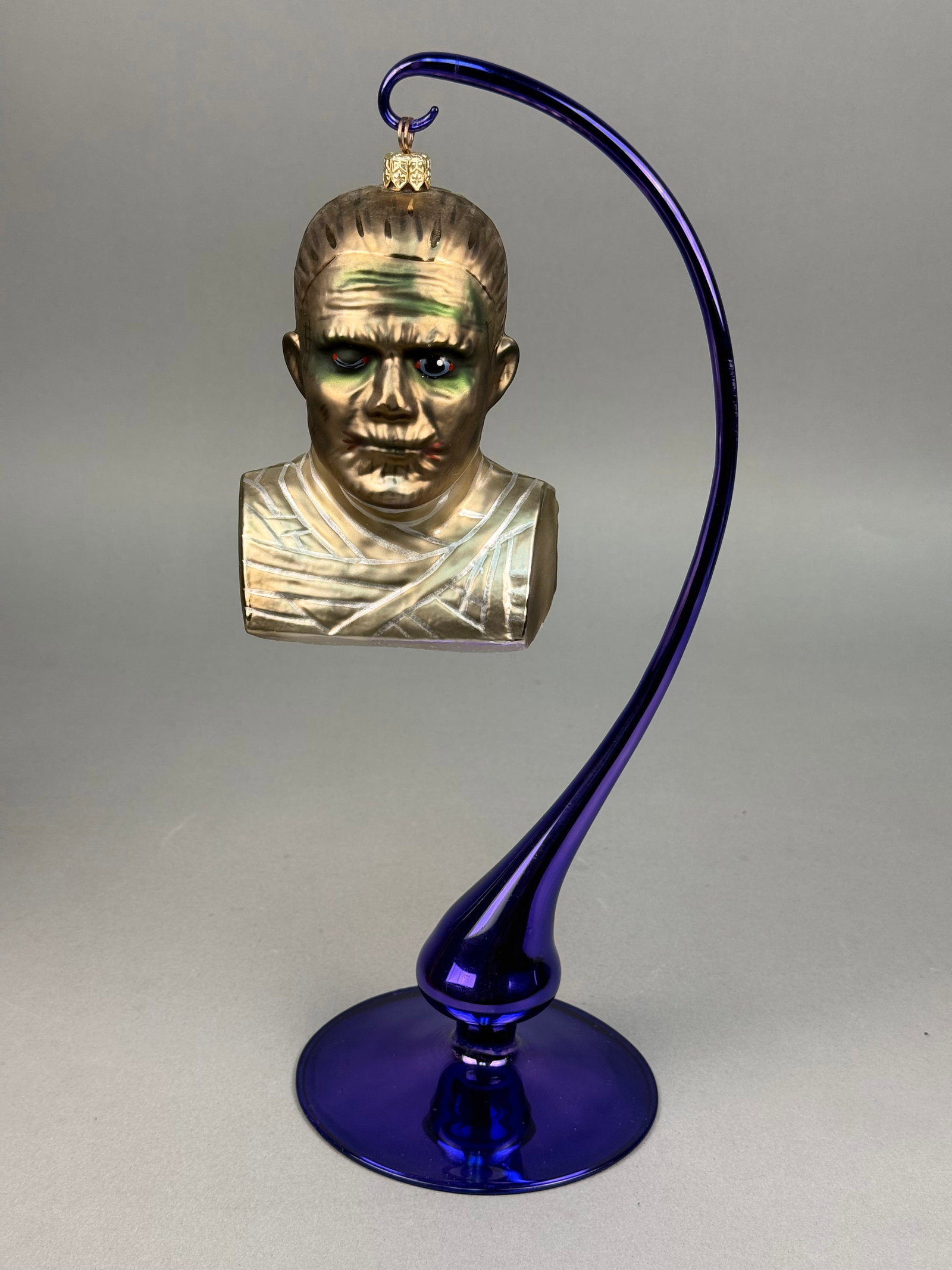 Radko Universal Studios The Mummy Blown Glass Ornament: A mouth-blown glass ornament by Christopher Radko depicting a bust of The Mummy from the Universal Classic Monsters series. The piece is finished in metallic gold and bronze tones, accented with hand-