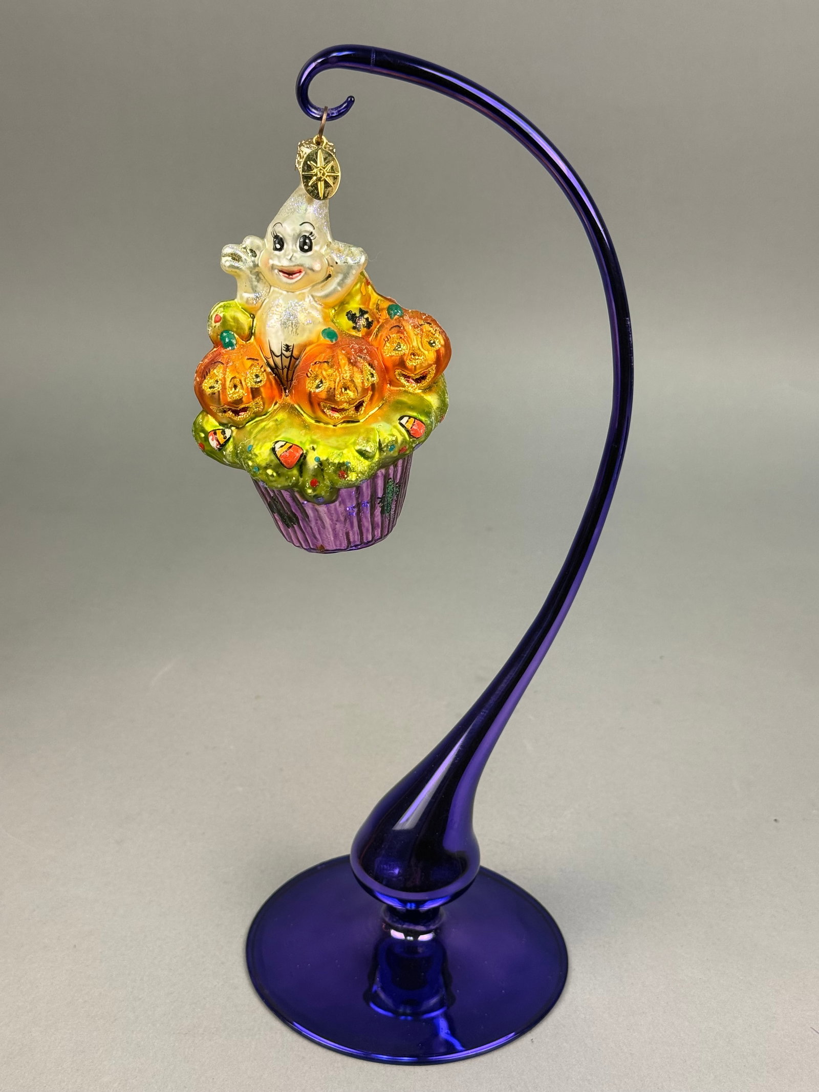 Radko Hand Blown Glass Ghostly Halloween Cupcake Ornament: A hand-blown glass holiday ornament featuring a whimsical ghostly cupcake design. The ornament depicts a white-glittered ghost with a smiling face and large black eyes emerging from behind three orang