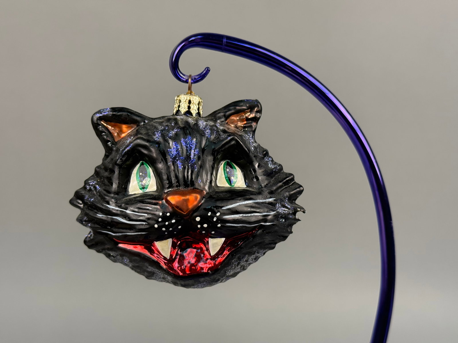 Radko Witchy Whiskers Black Cat Glass Halloween Ornament: A hand-painted mouth-blown glass holiday ornament manufactured by Christopher Radko. The piece, titled Witchy Whiskers, depicts a stylized black cat head with a textured surface designed to resemble r