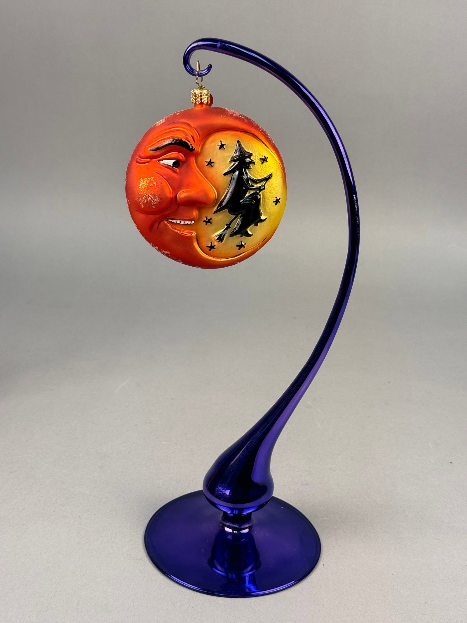 Radko Double Sided Halloween Man in the Moon Glass Ornament: A hand-blown and hand-painted glass Halloween ornament attributed to Christopher Radko, accompanied by a cobalt blue glass display stand. The double-sided, circular ornament features a frosted orange 