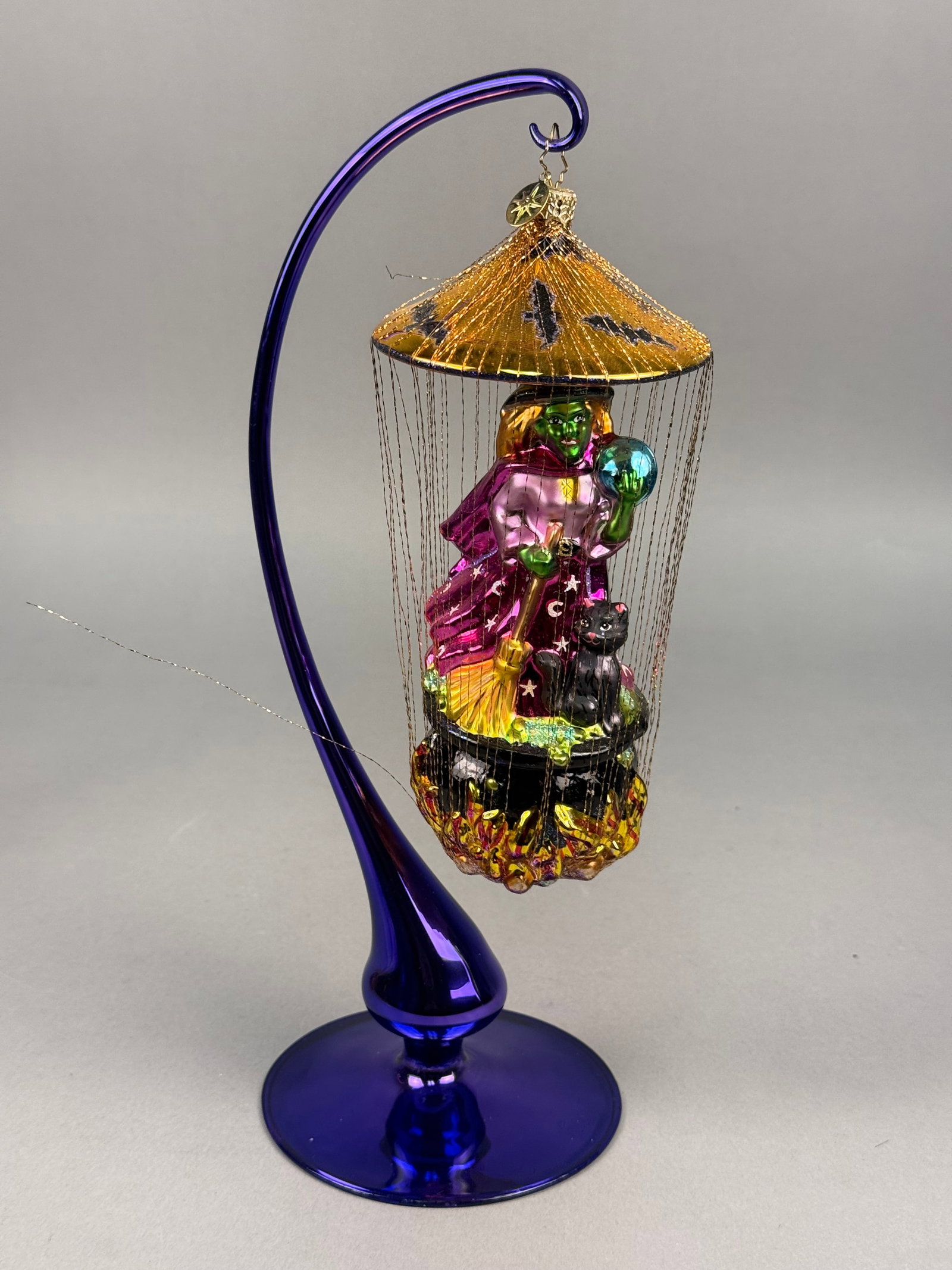 Radko Caged Halloween Witch Glass Ornament: A hand-blown glass holiday ornament by Christopher Radko depicting a Halloween scene. The central figure is a green-skinned witch dressed in a metallic magenta robe with silver star and crescent moon 