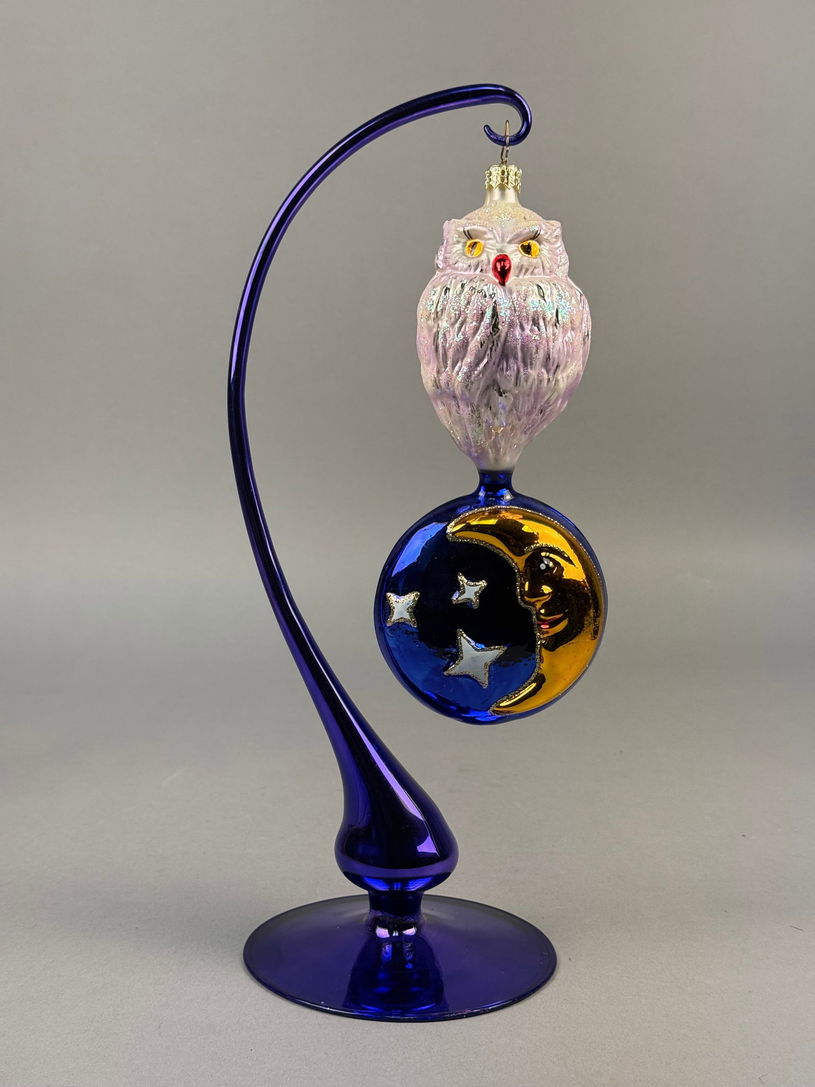Radko Hand-Blown Glass Owl and Moon Celestial Ornament: This hand-blown glass ornament from Christopher Radko features an owl and celestial motif. The piece consists of two integrated glass components. The upper section is a molded owl head in iridescent p