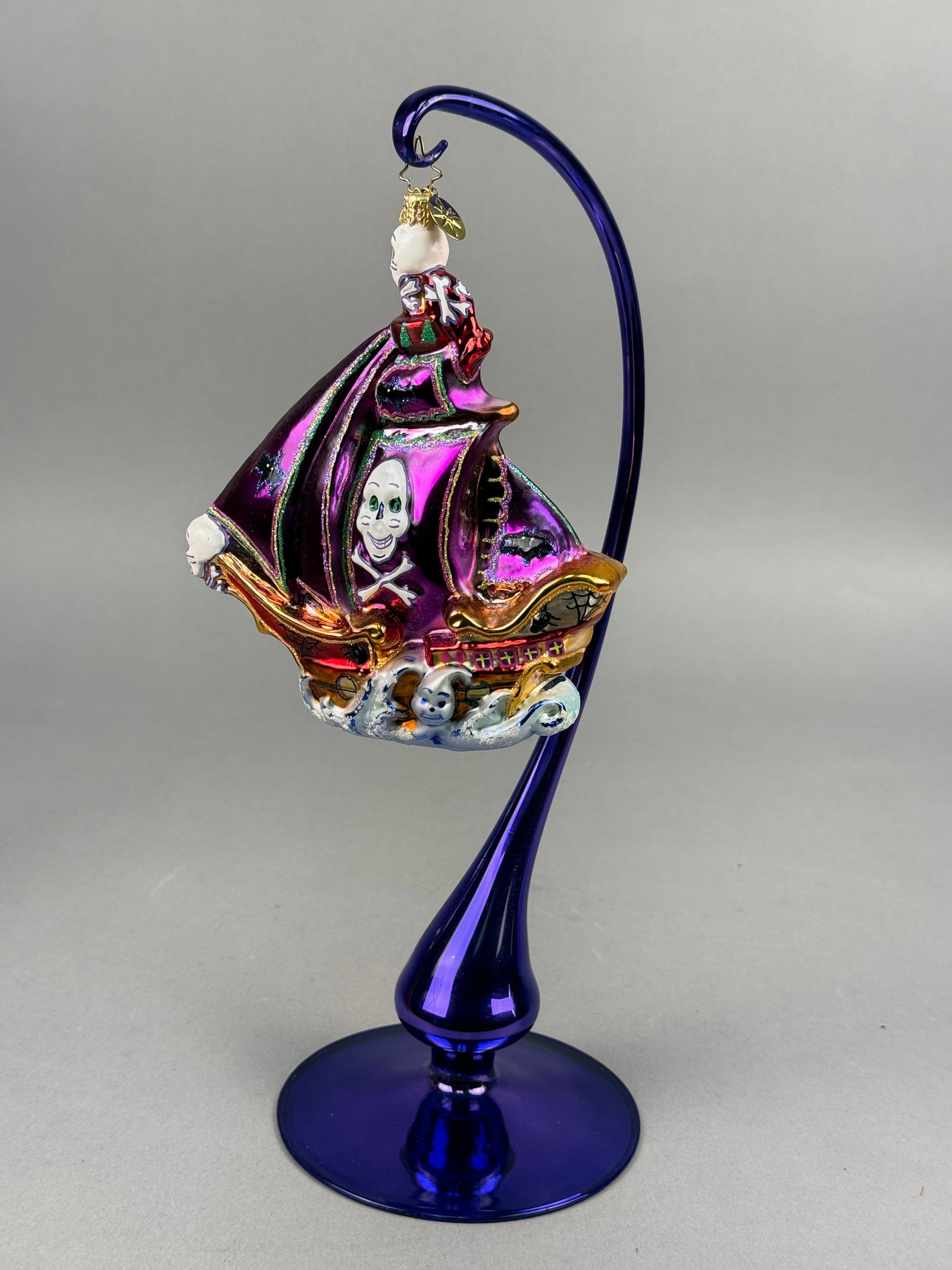 Radko Ghost Ship Halloween Glass Ornament: A mouth-blown glass Halloween ornament by Christopher Radko, titled Ghost Ship. The ornament is designed as a pirate ship featuring iridescent metallic purple sails and a hull accented with vibrant go