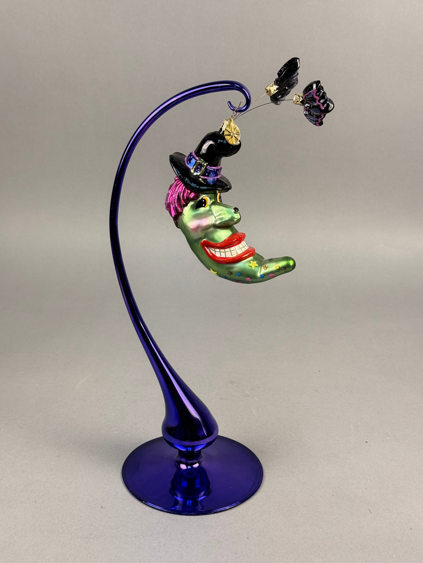 Radko Blown Glass Halloween Moon Ornament: The ornament depicts a green crescent moon face with a whimsical expression, red lips, and a toothy grin. The figure features pink glittered hair and wears a black witch hat with a purple glittered ba