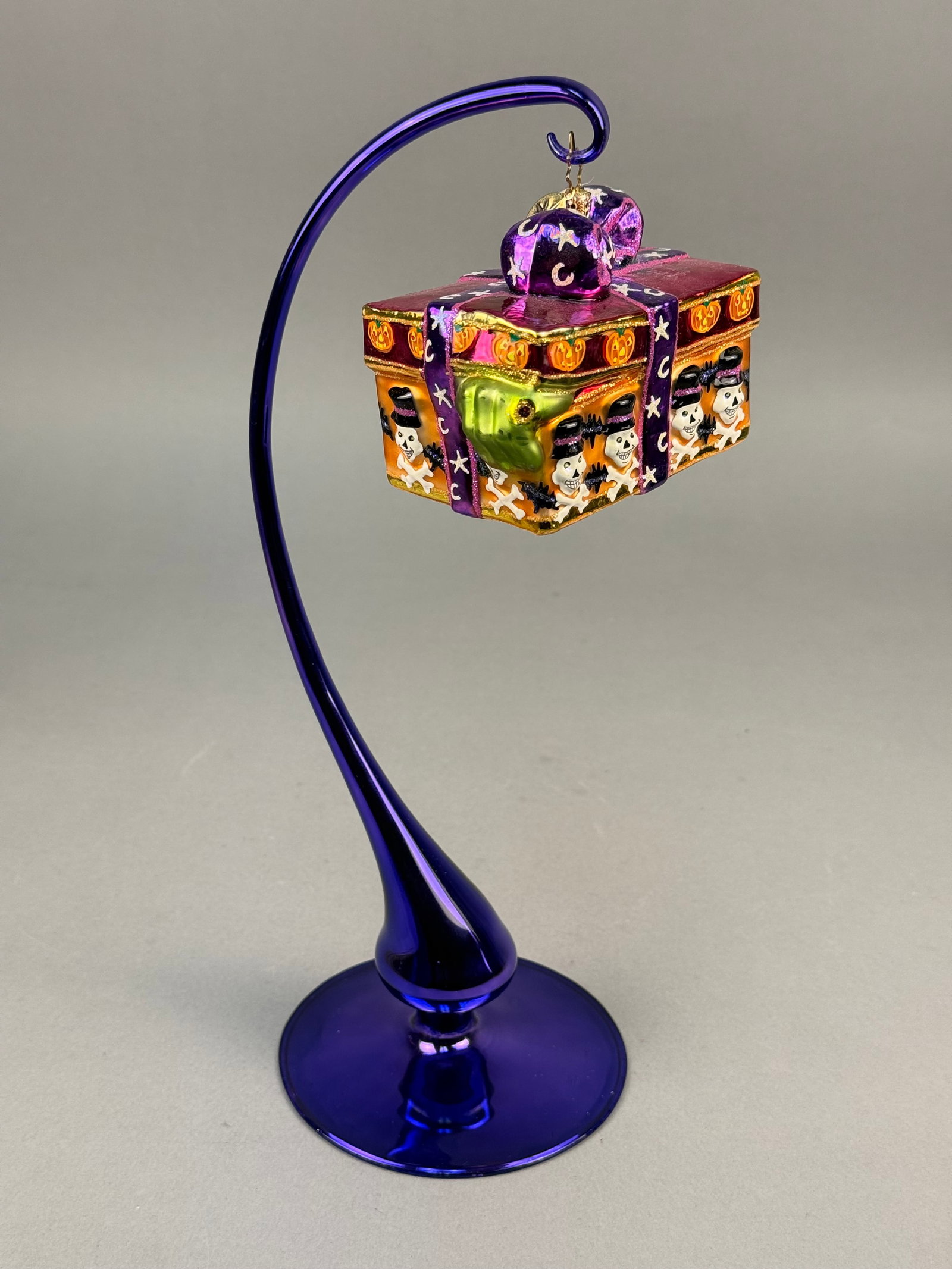 Radko Blown Glass Halloween Ornament: This lot consists of a hand-blown glass Halloween ornament by Christopher Radko. The ornament is designed as a rectangular gift box with an orange body and a deep purple lid. It retains the maker's si