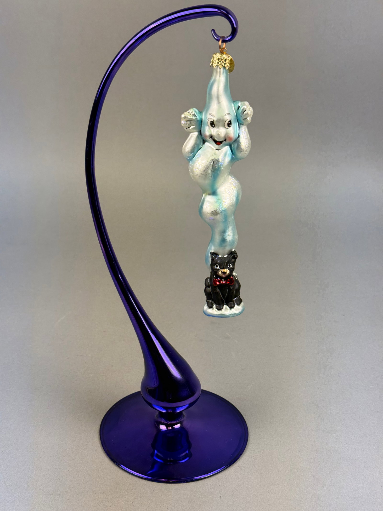 Radko Ghost and Black Cat Glass Ornament: The piece is designed as an elongated, iridescent light blue and white ghost with a hand-painted smiling face. The ghost is depicted holding a small black cat with a red bow tie at the end of its trai