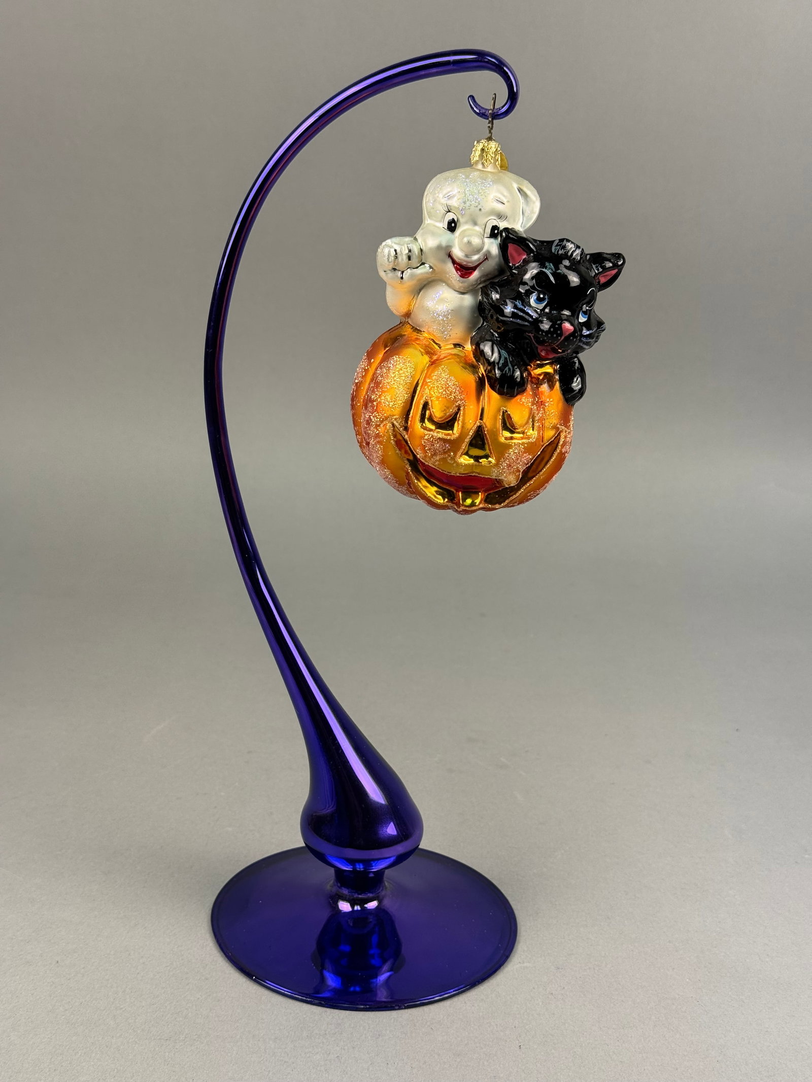Radko Blown Glass Halloween Ghost Cat and Pumpkin Ornament: A hand blown glass Halloween ornament by Christopher Radko, featuring a three-part composition consisting of a smiling white ghost, a black cat with blue eyes, and an orange jack-o-lantern. It is fitt