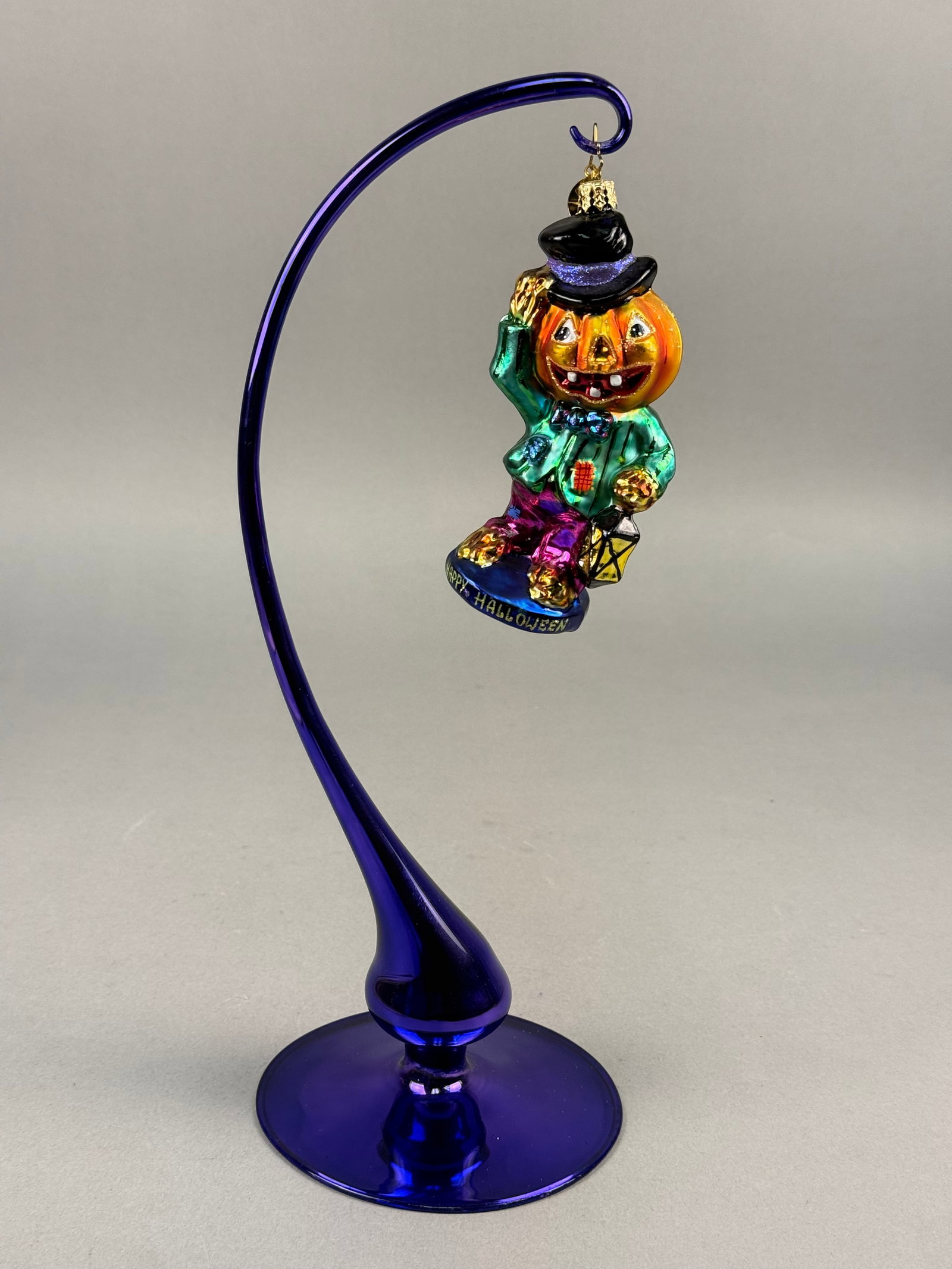 Radko Happy Halloween Blown Glass Ornament: A Christopher Radko hand-painted blown glass ornament featuring a whimsical Halloween figure. The character is designed with a jack-o-lantern head wearing a black top hat with a purple glittered band.