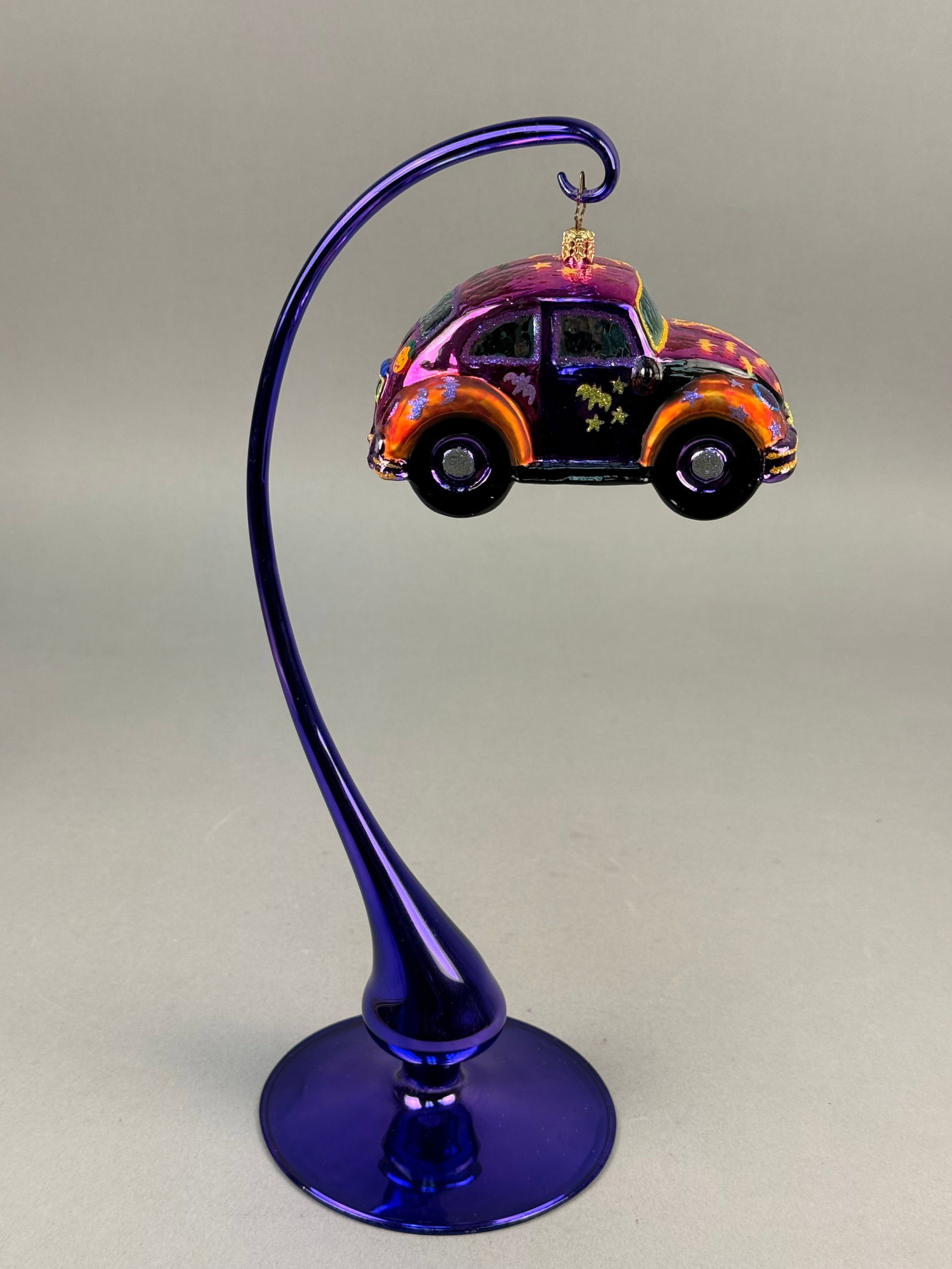 Christopher Radko Halloween-Themed Volkswagen Beetle Glass Ornament with Stand: A hand-blown glass holiday ornament by Christopher Radko, shaped like a classic Volkswagen Beetle with a Halloween motif. The piece features a multi-colored iridescent finish in shades of purple, oran