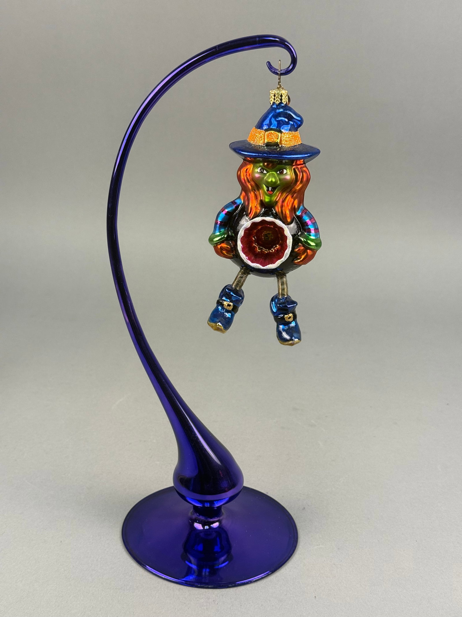 Christopher Radko Hand-Painted Glass Witch Cauldron Ornament and Blue Glass Stand: This lot features a hand-painted, mouth-blown glass hanging ornament by Christopher Radko, depicting a whimsical witch figure. The character is designed with a vibrant green face, large eyes, and a sm
