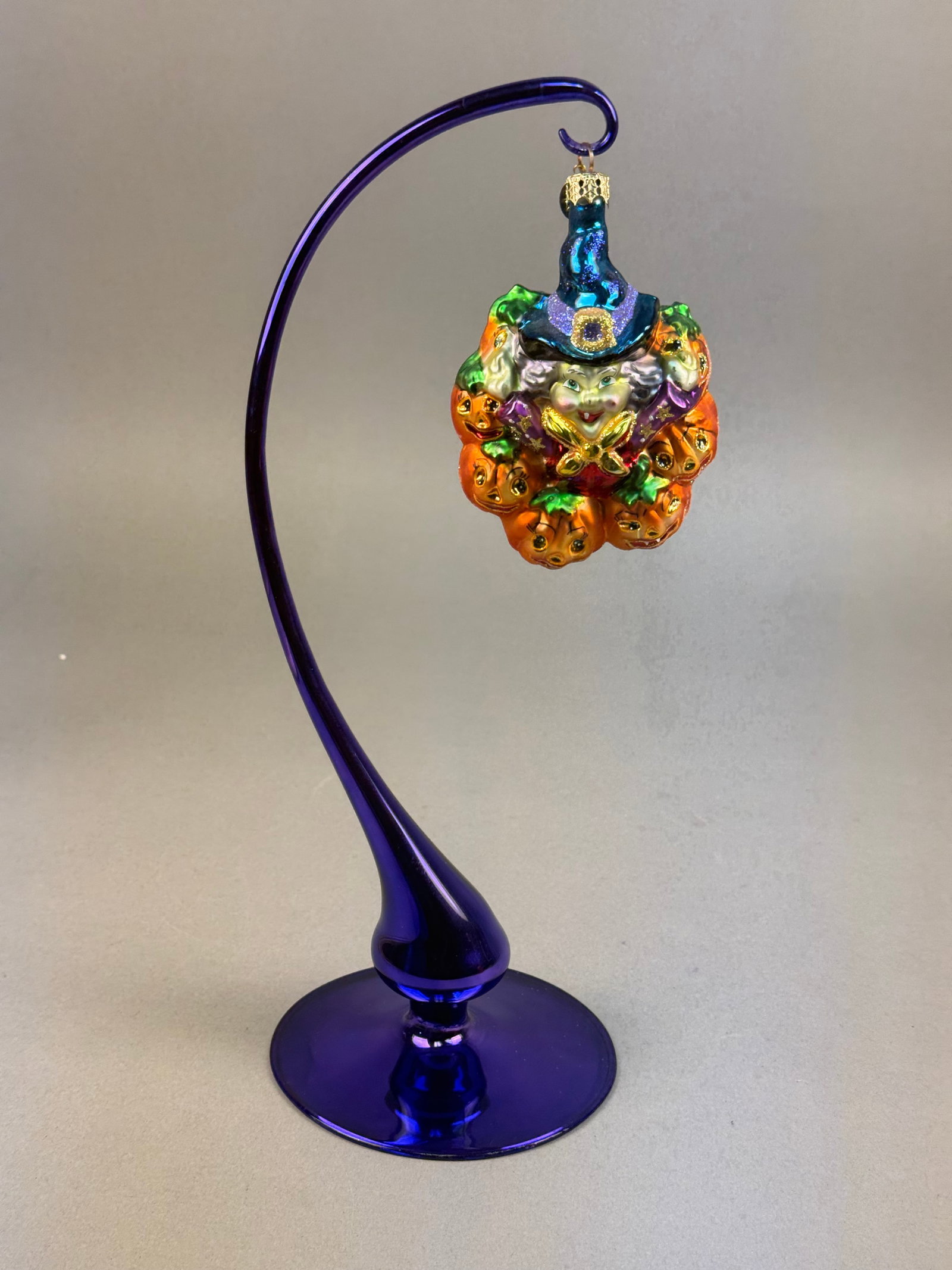 Christopher Radko Halloween Witch and Jack-O-Lantern Glass Ornament with Cobalt Blue Glass Stand: A hand-blown glass Halloween ornament produced by Christopher Radko. The ornament features a central witch figure with green eyes and a smiling expression, wearing a tall, curved, iridescent blue hat 