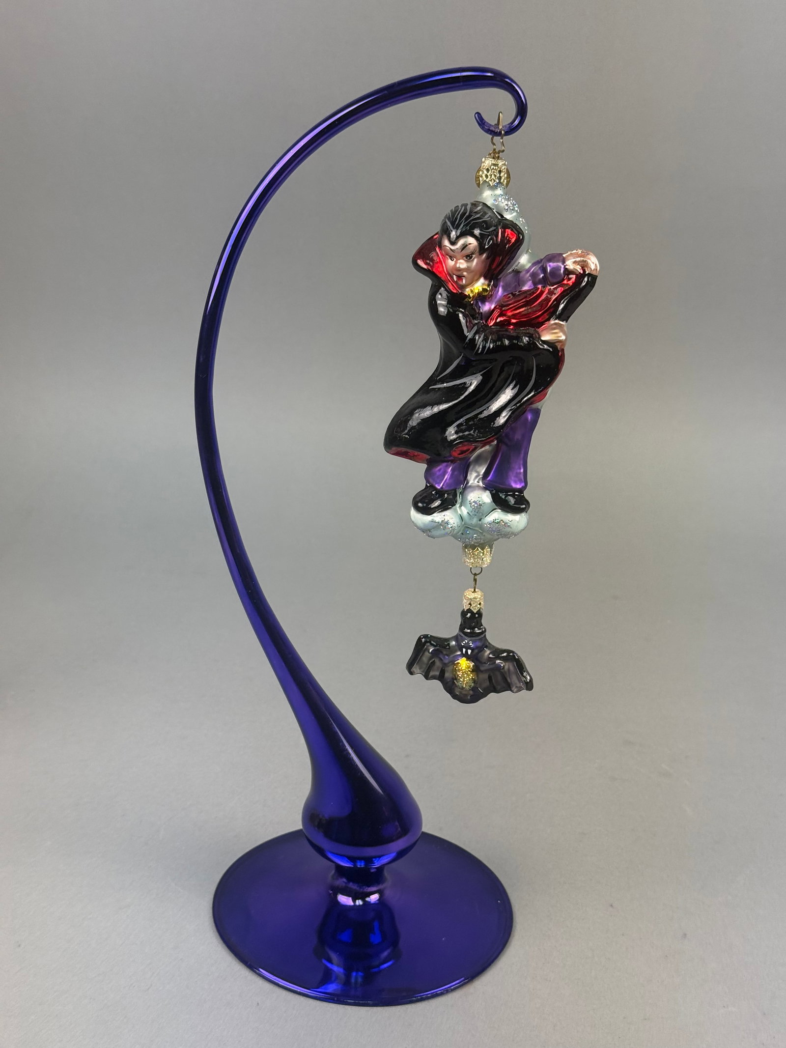 Christopher Radko Hand-Painted Blown Glass Vampire and Bat Halloween Ornament: A hand-painted blown glass holiday ornament produced by Christopher Radko, depicting a stylized vampire in a horizontal flying pose. The figure is dressed in a black cape with red lining, purple trous