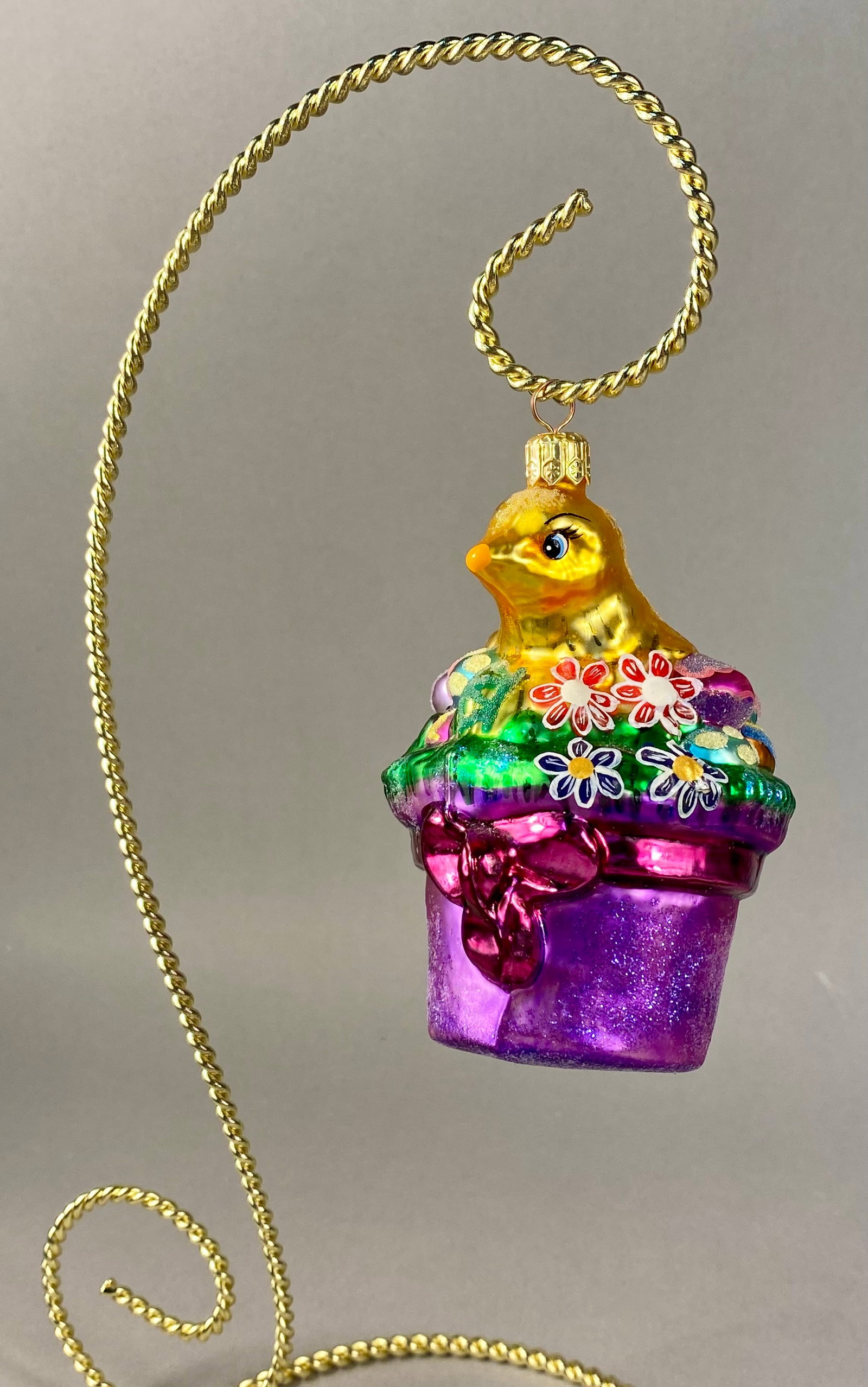 Hand-Blown Glass Easter Chick in Flower Pot Ornament: A mouth-blown glass holiday ornament featuring a yellow chick seated within a decorated flower pot. The chick is finished in a metallic yellow with blue eyes and an orange beak, accented by a frosted 
