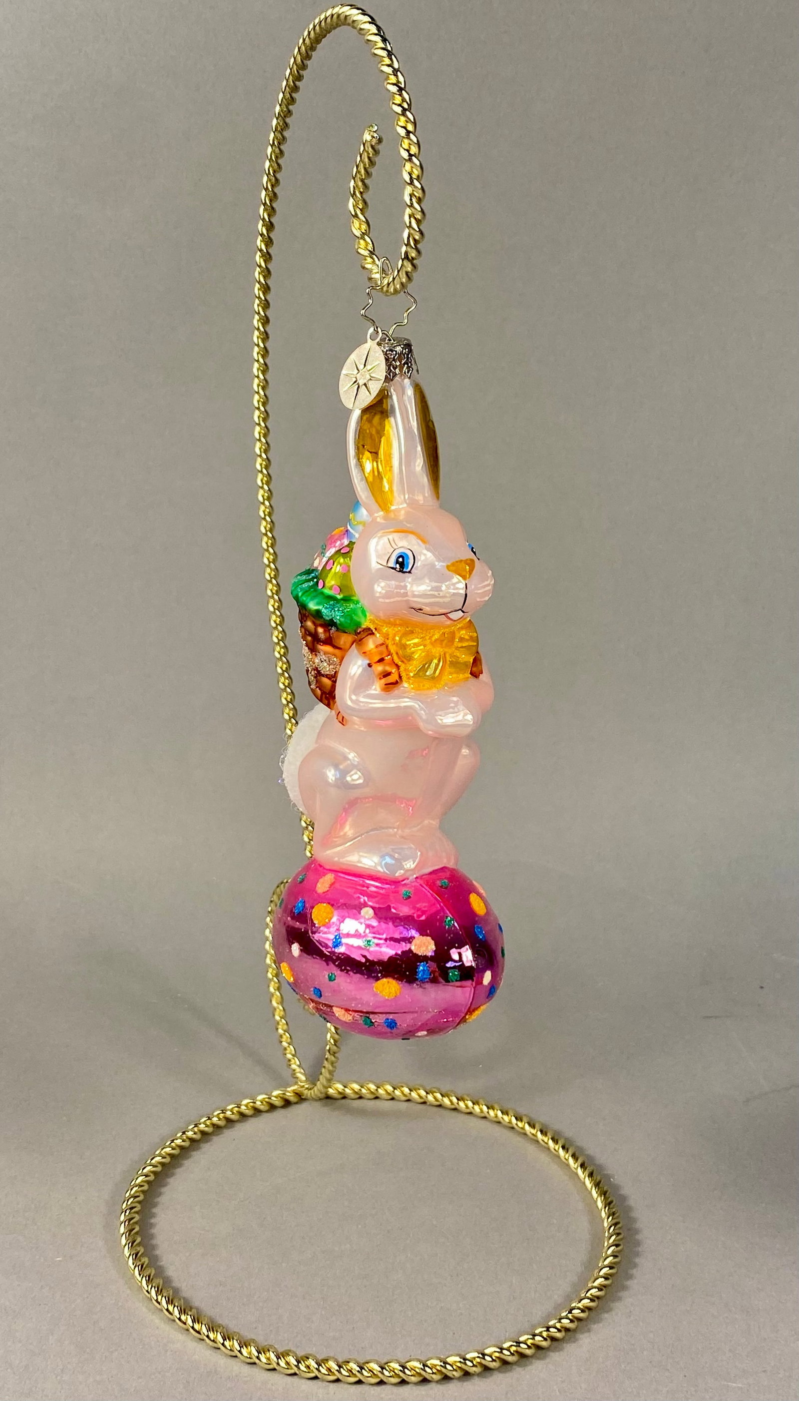 Christopher Radko Hoppy Holidays Hand Blown Glass Easter Bunny Ornament: A hand-painted, mouth-blown glass Easter ornament produced by Christopher Radko. The figurine depicts a light pink opalescent bunny standing upright atop a large fuchsia-pink Easter egg. The egg is de
