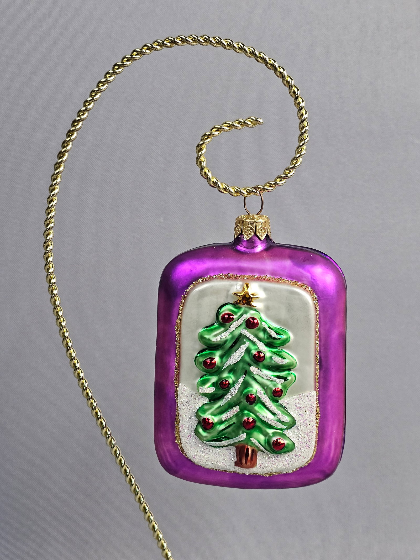 Christopher Radko Mouth-Blown Glass Christmas Ornament: This mouth-blown glass Christmas ornament by Christopher Radko is designed in a rectangular pillow shape using translucent magenta glass with rounded corners. The obverse features a central inset pane