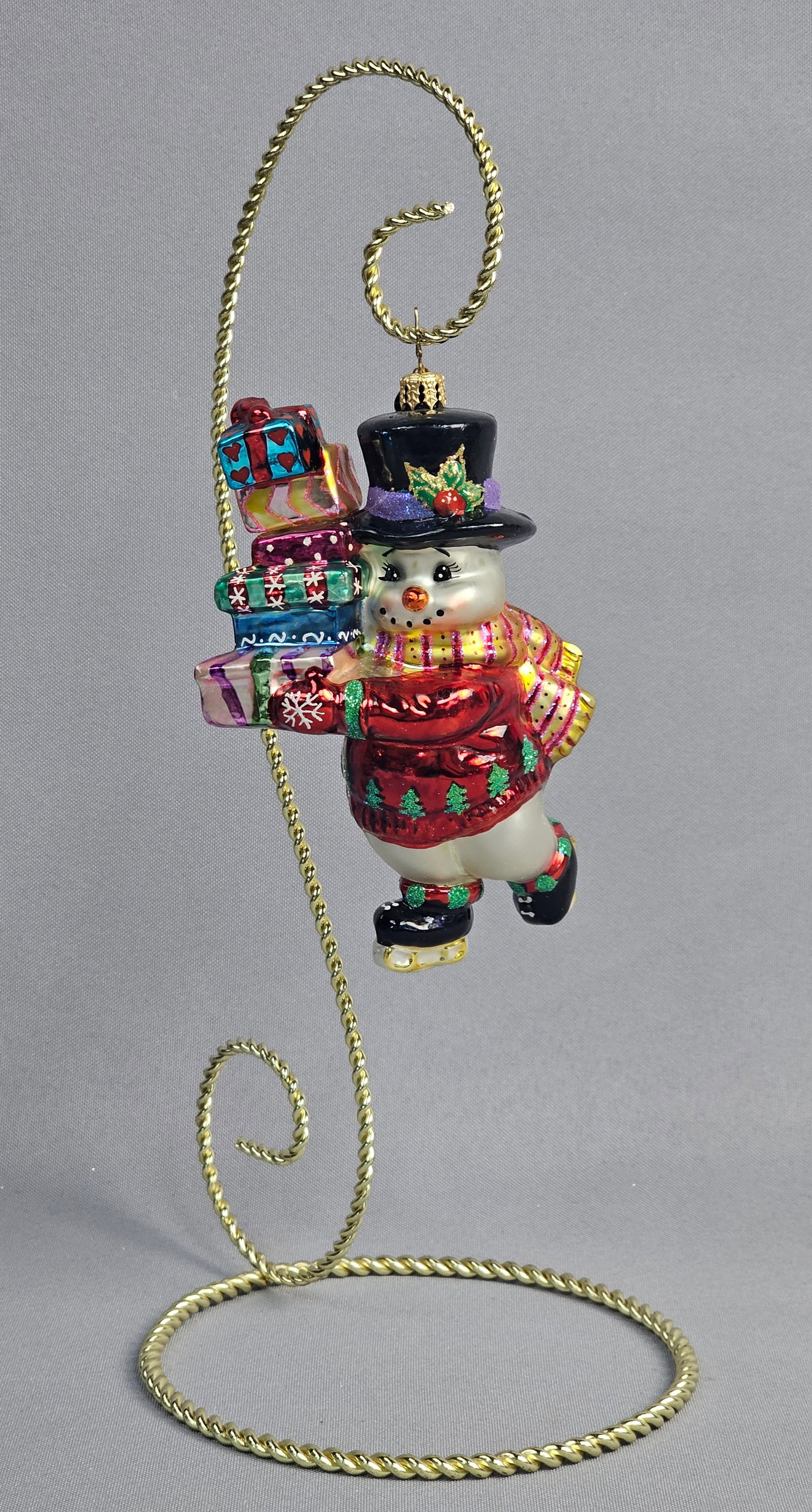 Christopher Radko Ice Skating Snowman with Gifts Glass Christmas Ornament: A Christopher Radko hand-painted mold-blown glass Christmas ornament featuring an ice-skating snowman carrying a stack of presents. The snowman is outfitted in a glossy red sweater decorated with gree