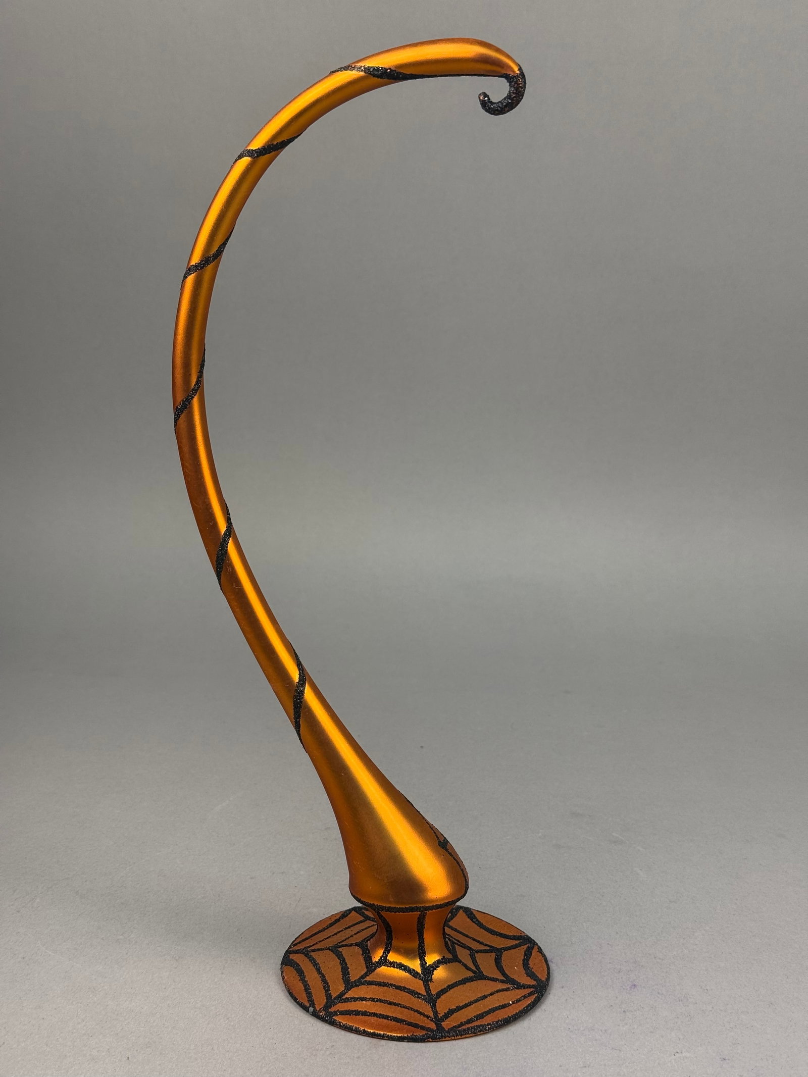 Contemporary Halloween Ornament Stand in Metallic Orange with Spider Web Motif: A contemporary decorative ornament display stand featuring a metallic orange finish and black glitter accents. The piece is composed of a flared circular base decorated with a spider web pattern, supp