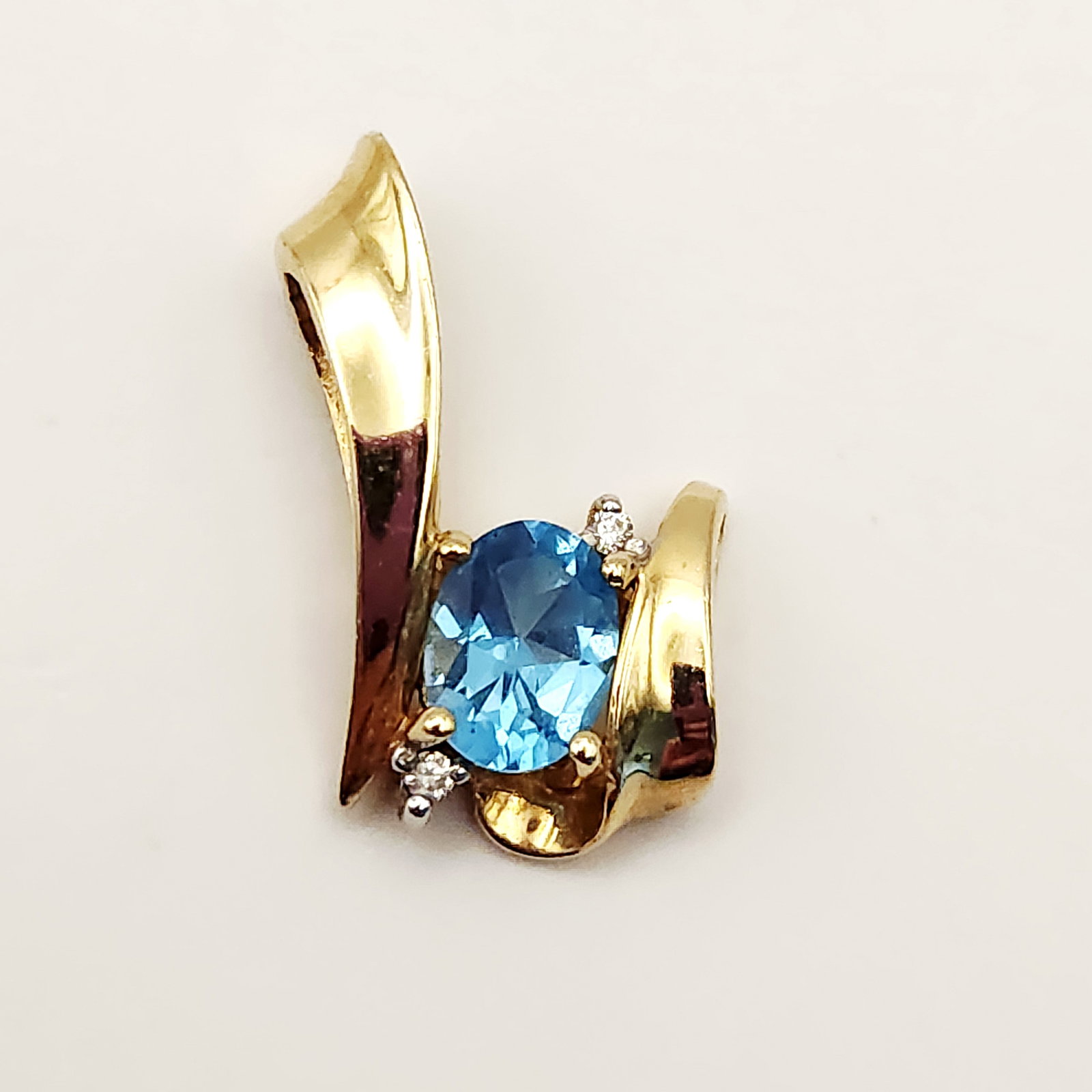 14K Gold Blue Topaz Pendant 3.2 Grams: A contemporary ribbon-style bypass pendant crafted in 14 karat yellow gold. The design features a centrally-set oval faceted light blue topaz & diamond accents. Total Weight: 3.2 grams Length: 0.75