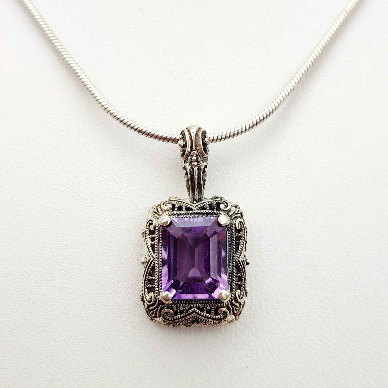 Sterling Amethyst Necklace 7.7 Grams: This sterling silver necklace features a rectangular pendant set with an emerald-cut faceted amethyst gemstone. The stone is mounted within an ornate sterling silver frame detailed with scrolling