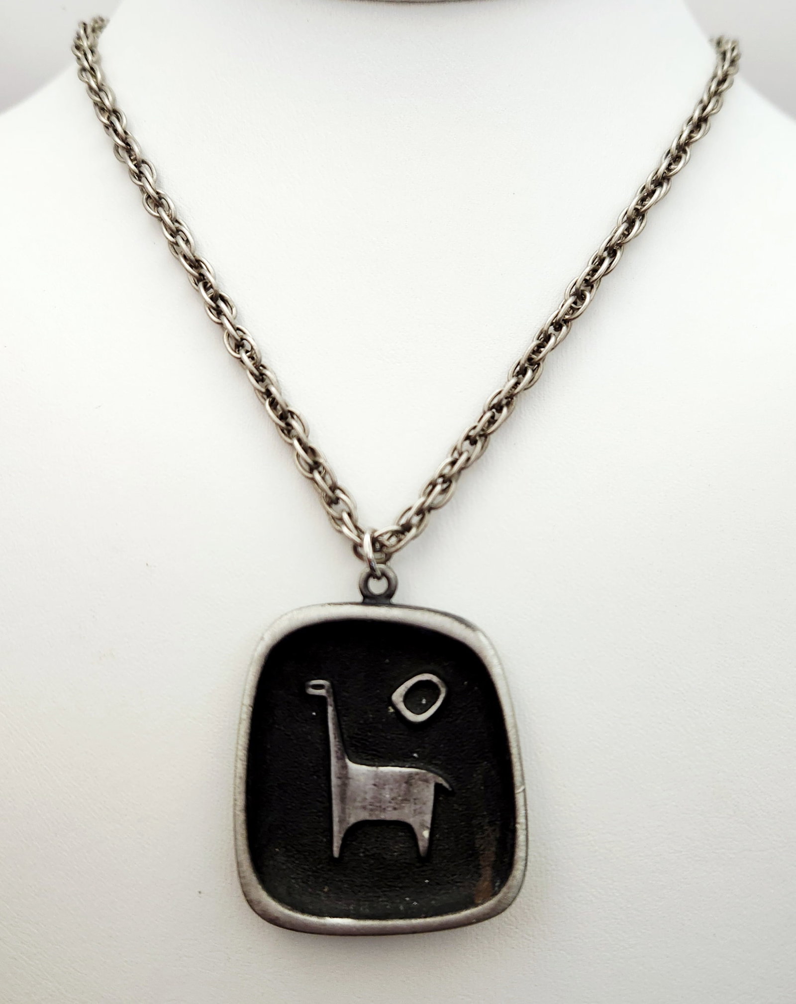 Mid-Century Modern Style Pewter Pendant Necklace with Stylized Animal Motif: A mid-century modern style necklace featuring a rounded rectangular pewter pendant. The front of the pendant depicts a raised, stylized long-necked animal figure standing beside an oval motif, both se