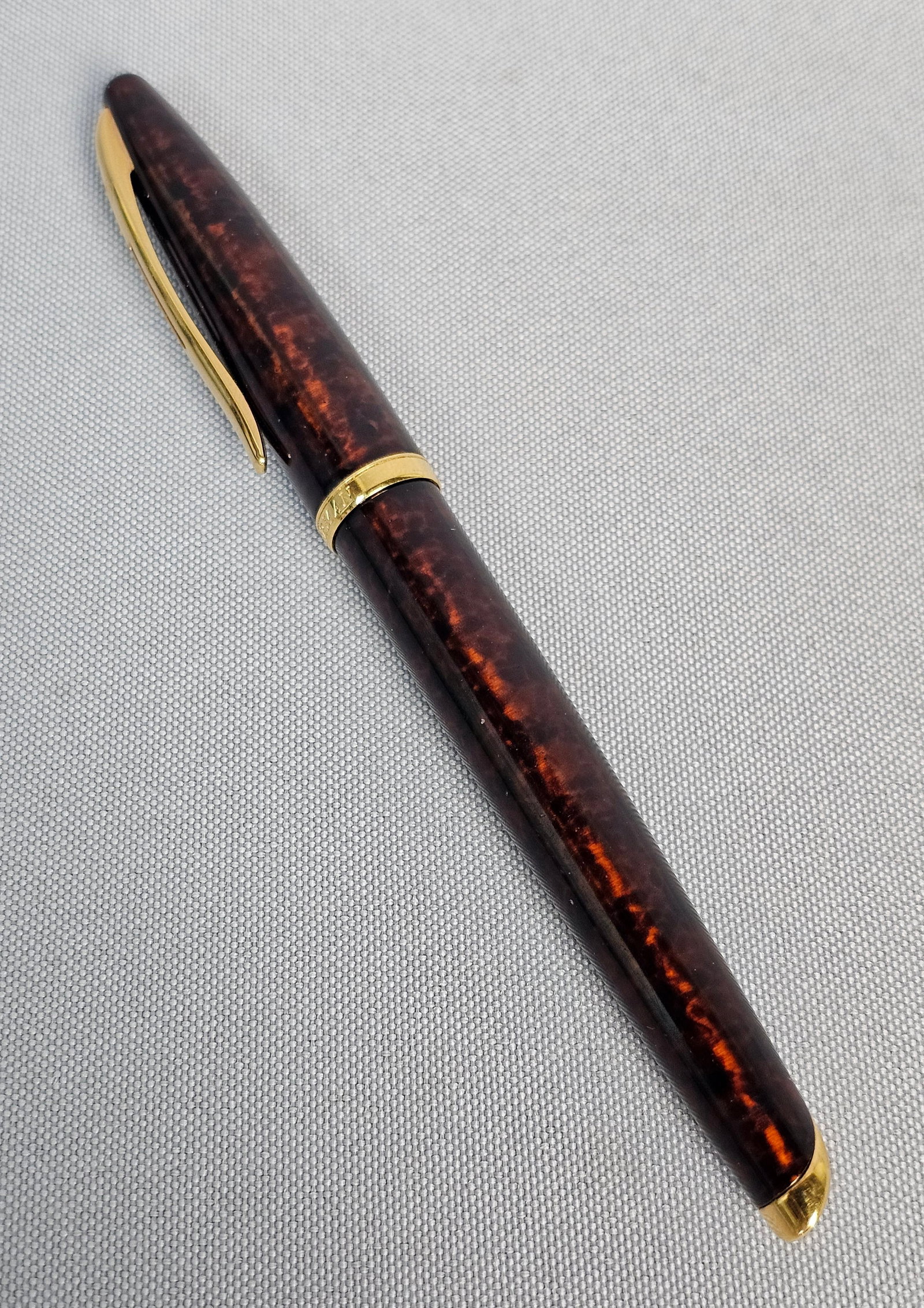 Waterman Carene Marine Amber Rollerball Pen: The body is finished in Marine Amber lacquer, a rich marbled pattern of deep brown and golden tones. The pen is accented with gold-plated trim, including the signature open-slotted clip and a tapered